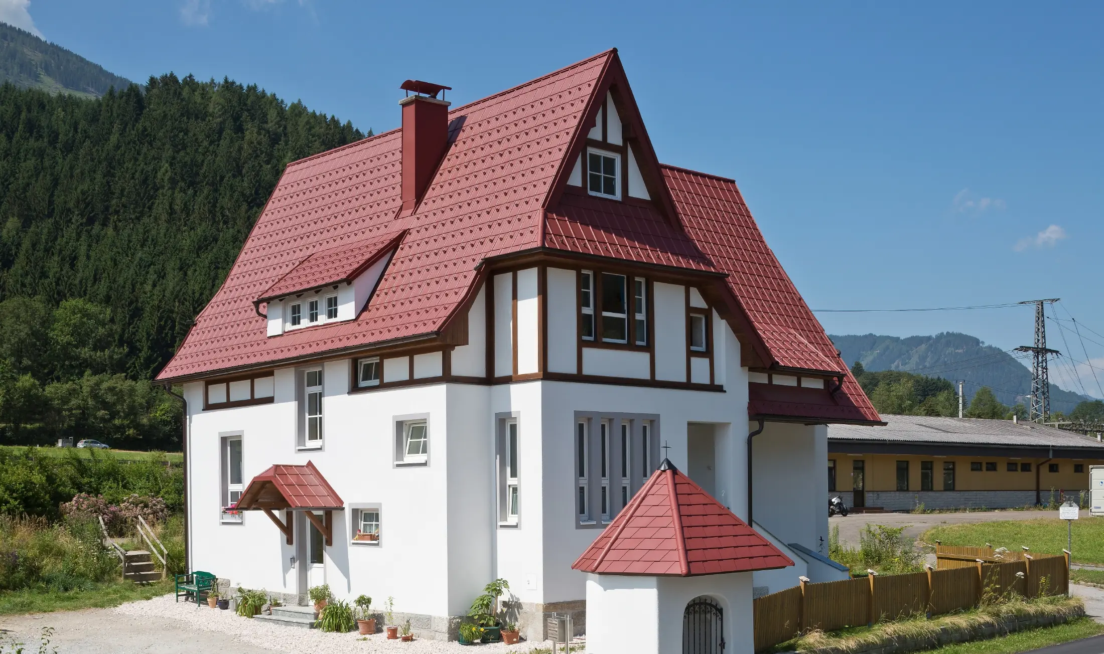 Šikmá strecha so Falcovanou škridlou PREFA v červenej farbe P.10 tmavočervená s mnohými detailmi, ako sú vikiere, vežičky a obruby komínov. Prístrešok a kaplnka pred domom sú tiež pokryté plechovou krytinou PREFA.