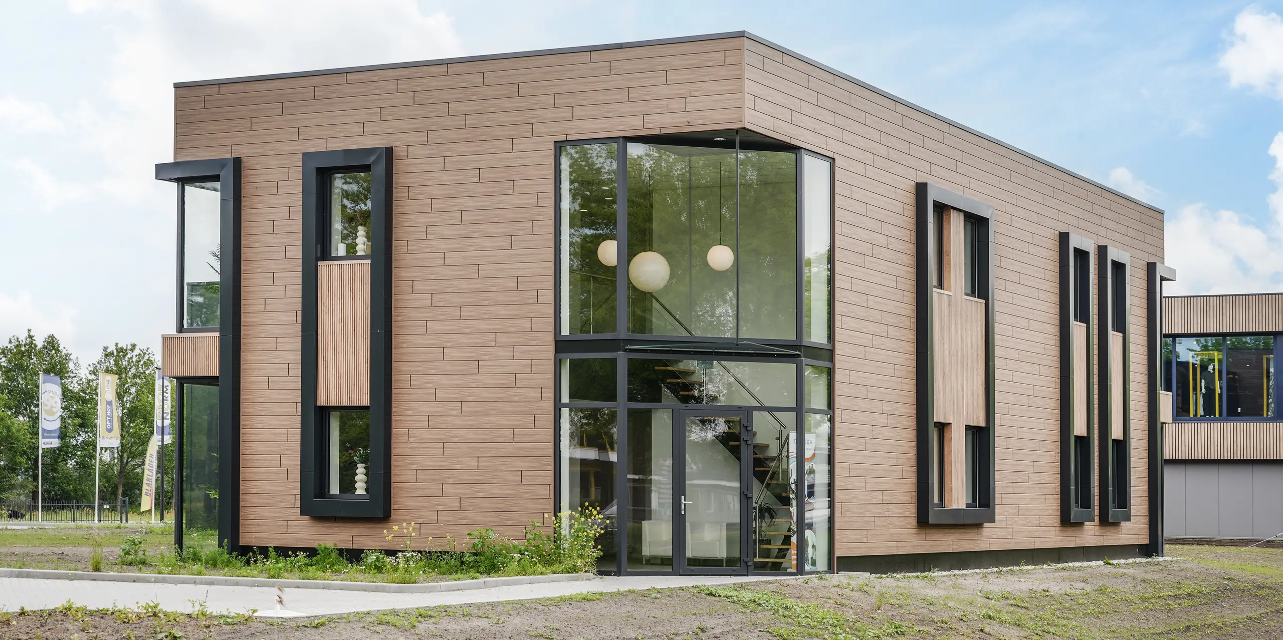Modernes Firmengebäude der Dokter Schoonmaakorganisatie in Wolvega mit markanter Fassadenarchitektur aus walnussbraunen PREFA Sidings. Großzügige Glasflächen, elegante schwarze Fensterrahmen und die natürliche Holzoptik setzen starke Akzente. Die hochwertige Aluminiumfassade überzeugt durch Langlebigkeit, Witterungsbeständigkeit, Korrosionsschutz und dauerhaft attraktive Optik bei minimalem Pflegeaufwand.