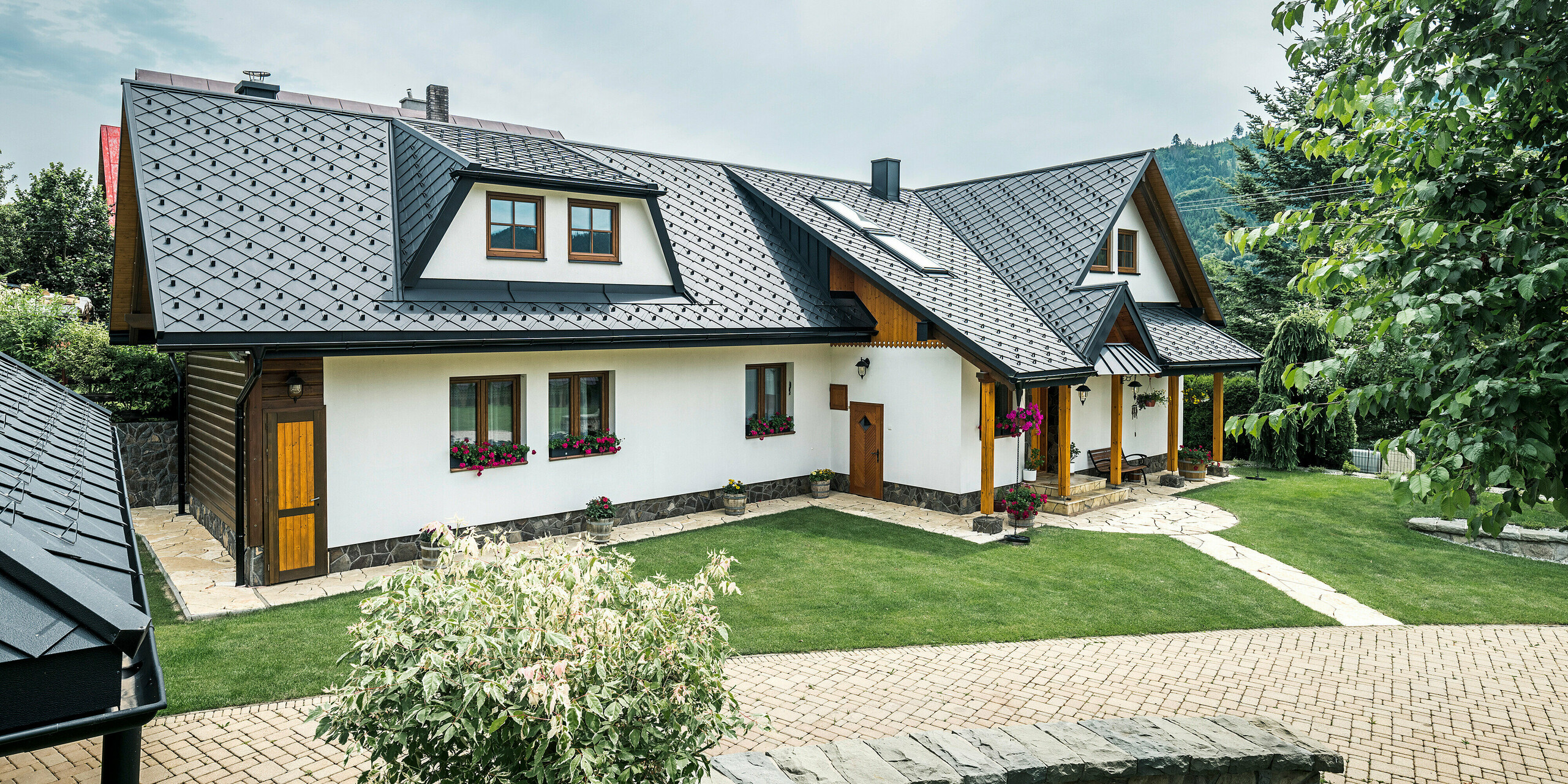 Pohľad na tradičný rodinný dom v Starej Bystrici na Slovensku so strechou zo strešných šablón 29 × 29 v P.10 čiernej farby. Hliníkové šablóny rovnomerne presahujú trapézový vikier v prednej časti budovy. V popredí snímky je vidieť udržiavanú záhradu. Vstupný priestor je chránený prístreškom oplášteným materiálom PREFALZ. Starostlivo navrhnutá príjazdová cesta a kvetinová výzdoba podčiarkujú prívetivý a štýlový celkový obraz.