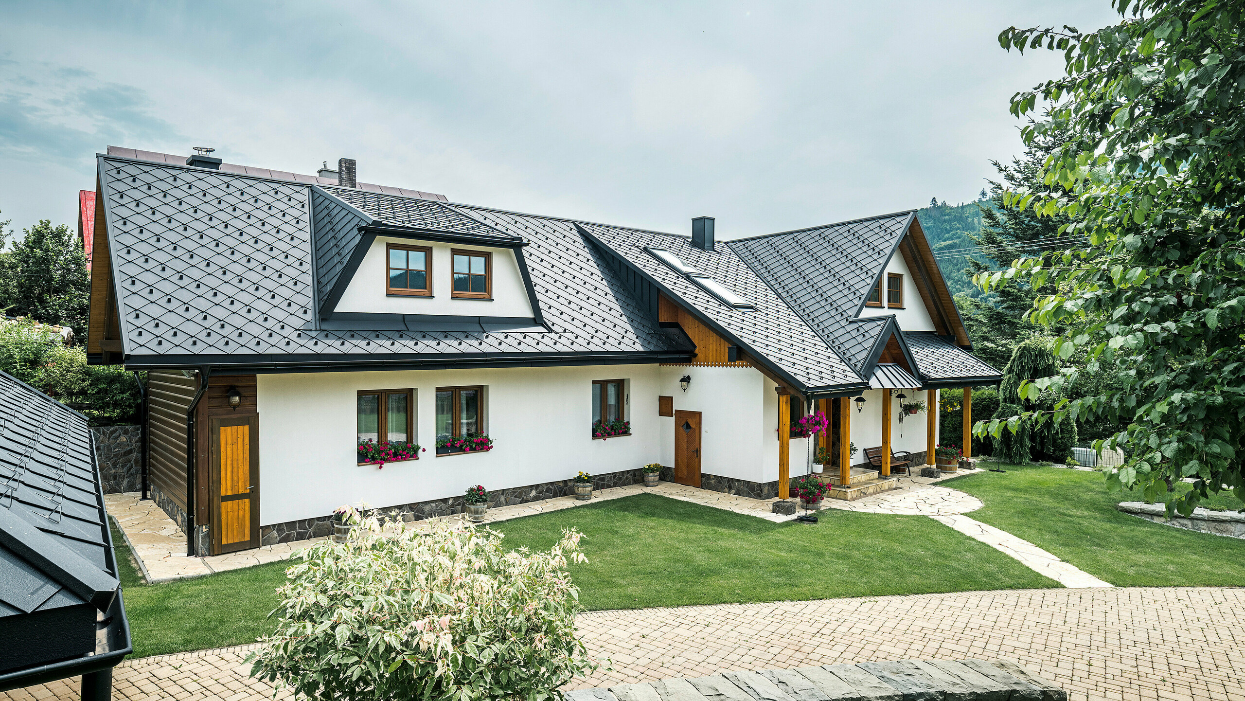 Pohľad na tradičný rodinný dom v Starej Bystrici na Slovensku so strechou zo strešných šablón 29 × 29 v P.10 čiernej farby. Hliníkové šablóny rovnomerne presahujú trapézový vikier v prednej časti budovy. V popredí snímky je vidieť udržiavanú záhradu. Vstupný priestor je chránený prístreškom oplášteným materiálom PREFALZ. Starostlivo navrhnutá príjazdová cesta a kvetinová výzdoba podčiarkujú prívetivý a štýlový celkový obraz.