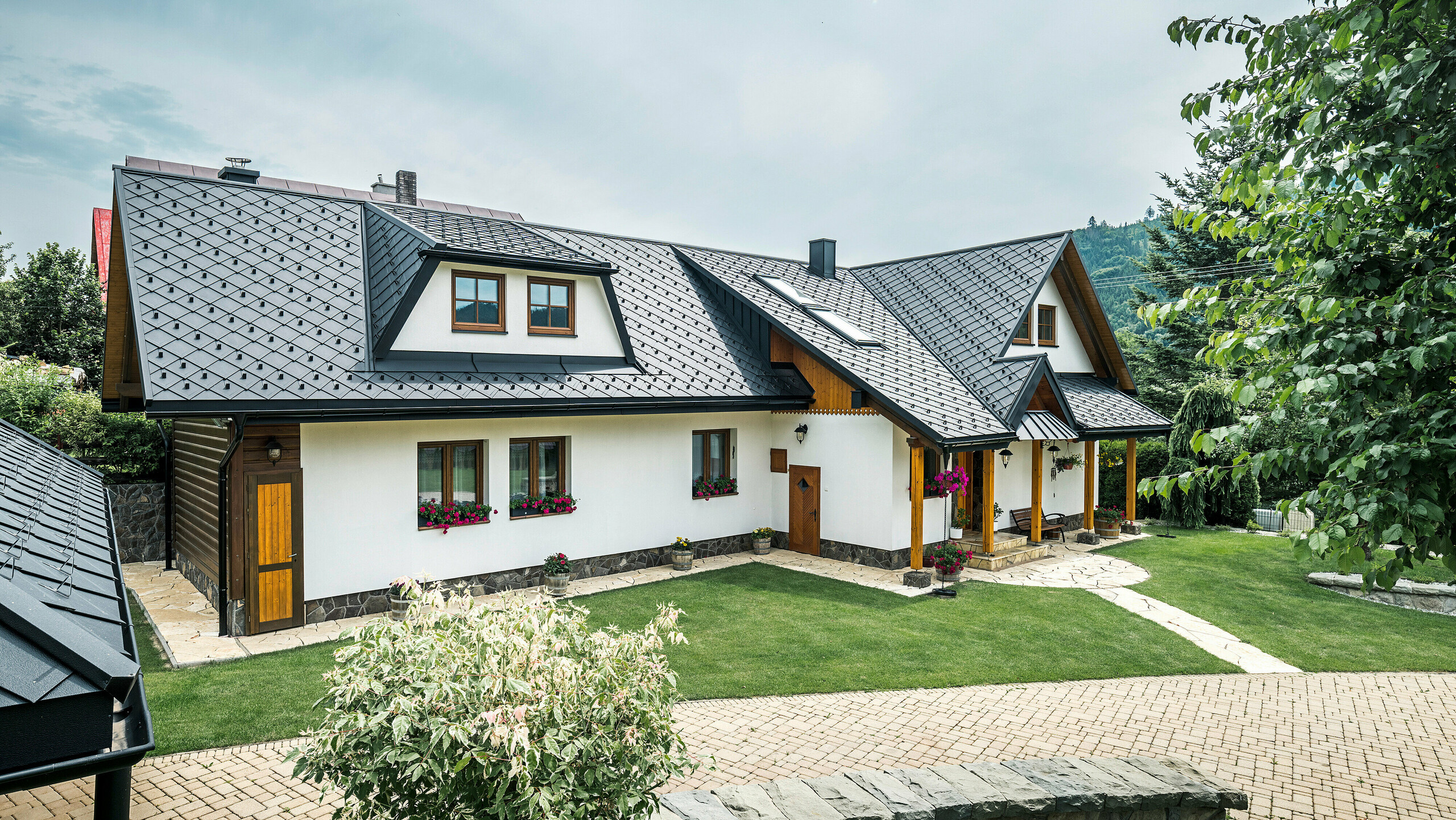 Pohľad na tradičný rodinný dom v Starej Bystrici na Slovensku so strechou zo strešných šablón 29 × 29 v P.10 čiernej farby. Hliníkové šablóny rovnomerne presahujú trapézový vikier v prednej časti budovy. V popredí snímky je vidieť udržiavanú záhradu. Vstupný priestor je chránený prístreškom oplášteným materiálom PREFALZ. Starostlivo navrhnutá príjazdová cesta a kvetinová výzdoba podčiarkujú prívetivý a štýlový celkový obraz.