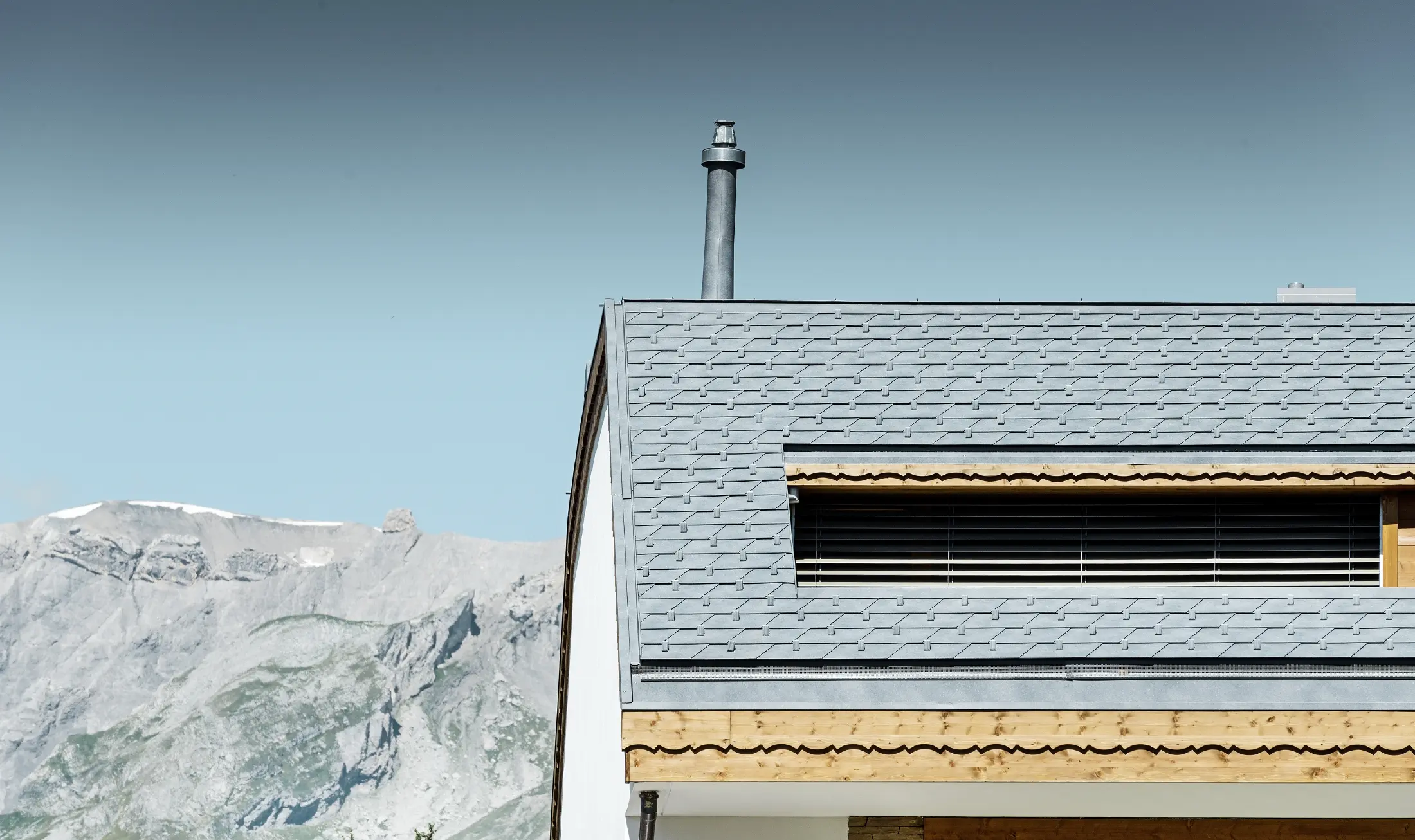 Bytový dom v Crans-Montane s horami v pozadí s fasádou s hravými drevenými prvkami a hliníkovou šindľovou strechou PREFA vo farbe kamenná šedá
