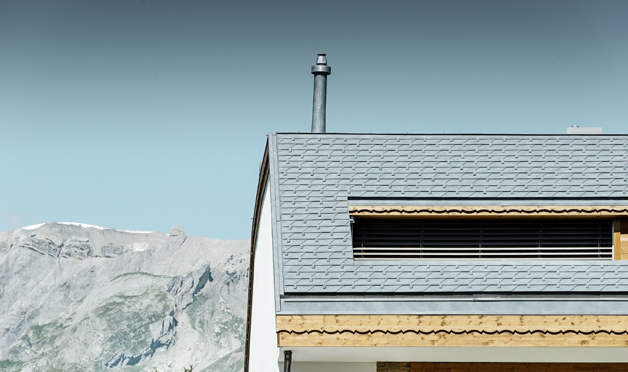 Bytový dom v Crans-Montane s horami v pozadí s fasádou s hravými drevenými prvkami a hliníkovou šindľovou strechou PREFA vo farbe kamenná šedá
