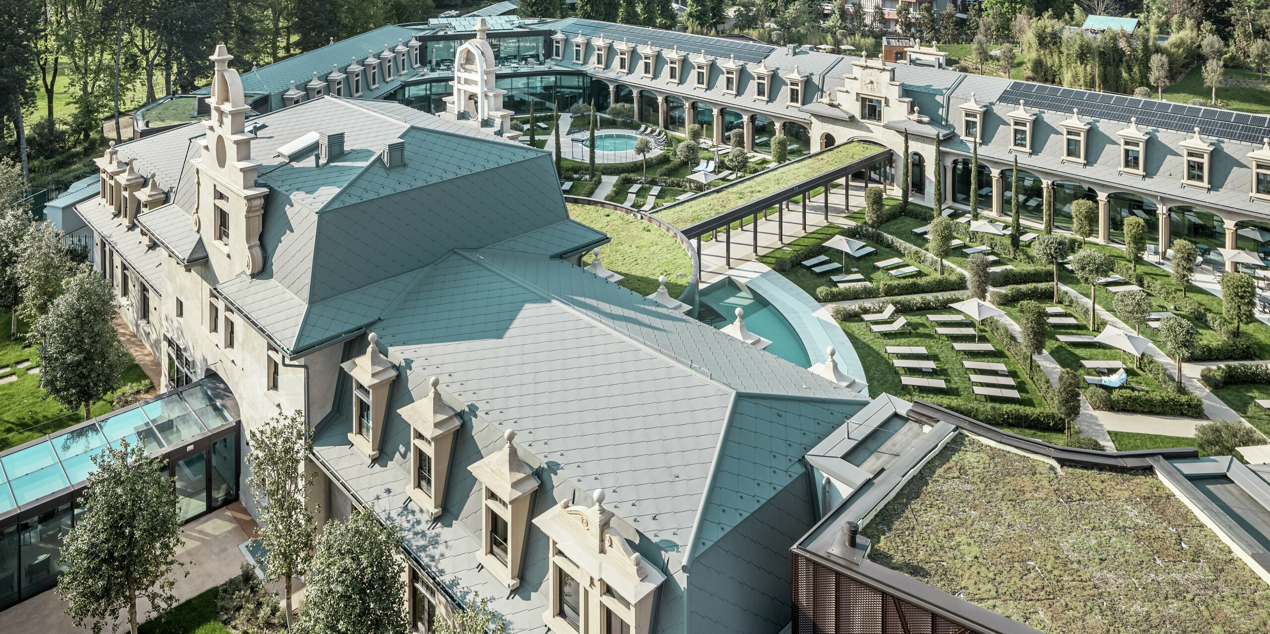 Panoramatický výhľad na kompletne zrekonštruovaný termálny komplex „De Montel – Terme Milano“ s rozľahlými záhradami, kľukatými cestičkami a oddychovými zónami okolo početných termálnych bazénov. Strechy sú pokryté strešnou šablónou PREFA 44 × 44 v odtieni P.10 svetlošedá, doplnenou o integrované fotovoltické moduly a zelené plochy. Historická architektúra a udržateľné materiály sa spájajú do inovatívneho mestského rekreačného priestoru.