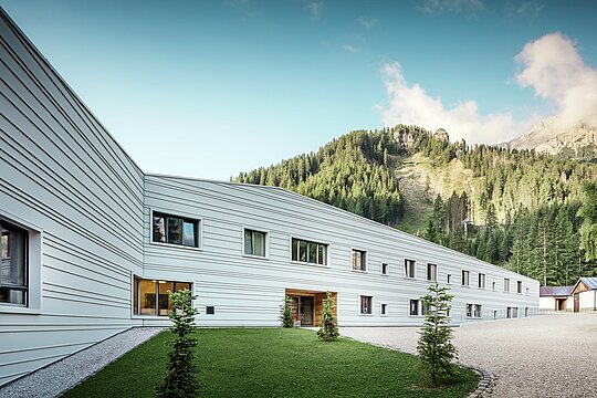 Hliník sa stretáva s precíznosťou – harmonický dialóg medzi technikou a prírodou. Pred impozantnou kulisou Dolomitov sa prezentuje „Villaggio Alpino Tempesti“ s jasnými líniami a moderným dizajnom. Fasáda z PREFALZ v farbe P.10 prefa biela je nielen mimoriadne odolná, ale vďaka svojej svetlej farbe tiež zabezpečuje príjemné mikroklíma, pretože odráža slnečné lúče. Zelené plochy a drevené prvky vo vstupnej časti vojenskej budovy vytvárajú prekvapivo príjemnú atmosféru.