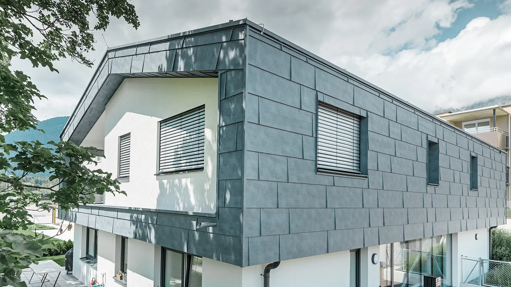 Moderný rodinný dom bez previsnutej strechy; horné poschodie bolo opláštené fasádnymi panelmi FX.12 od firmy PREFA vo farbe kamenná šedá, zvyšok fasády bol omietnutý nabielo.