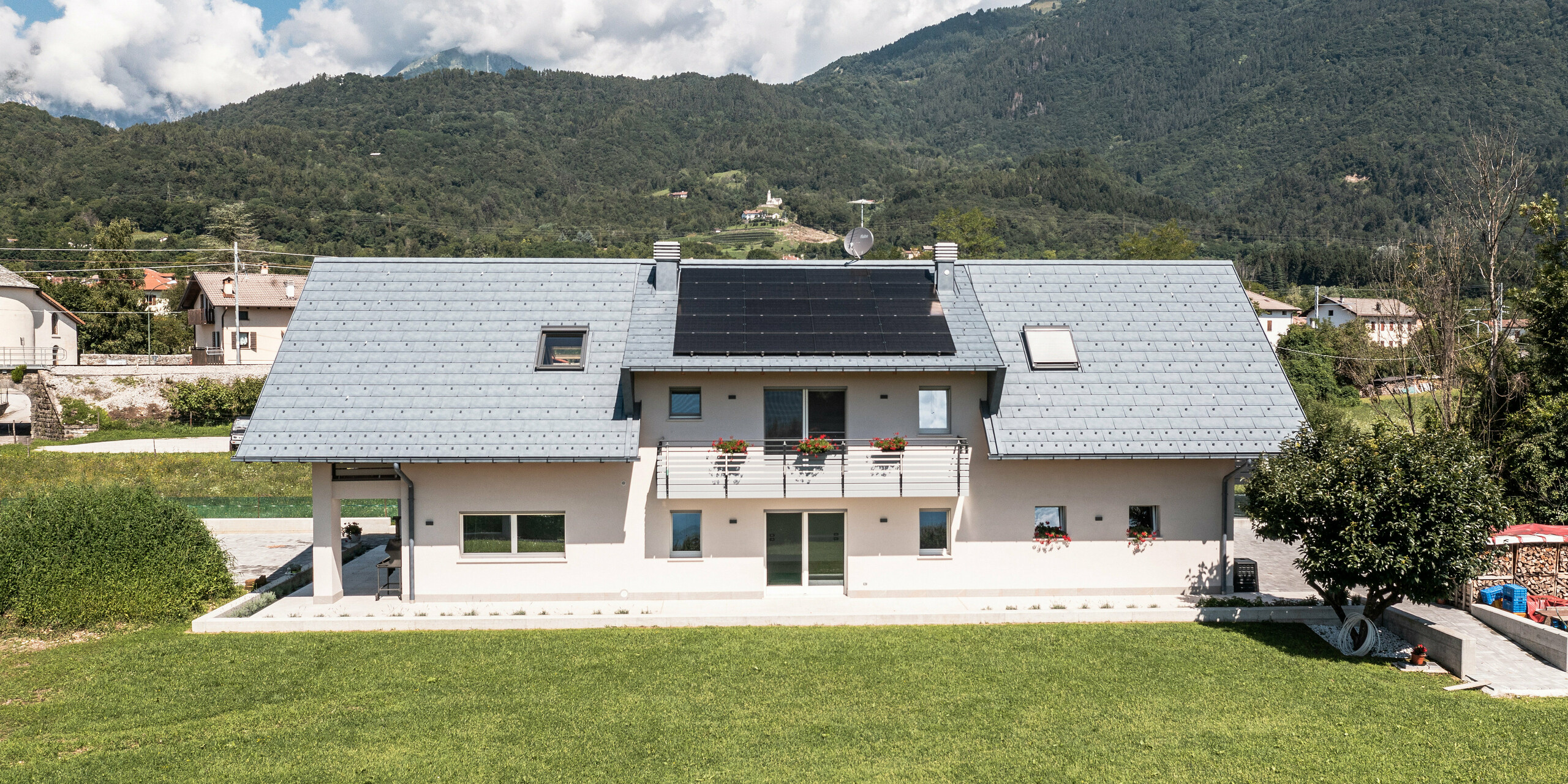 Idylický rodinný dom v Bellune, vybavený strešnými panelmi PREFA R.16 vo farbe antracit, umiestnený uprostred sviežej zelenej lúky. Čisté línie architektúry a moderná strecha vytvárajú nádherný kontrast k tradičným vidieckym domom v pozadí a okolitej horskej krajine.
