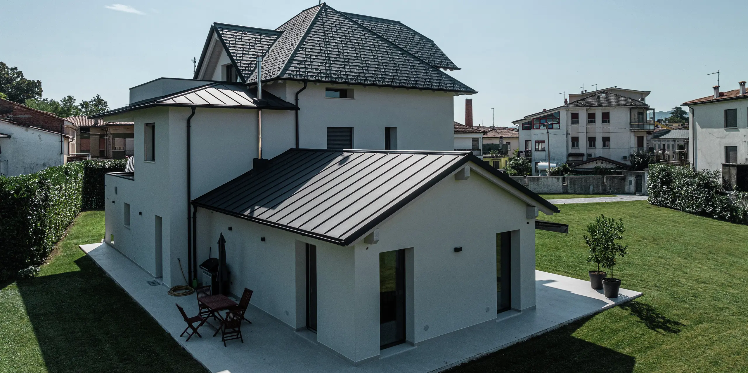 Pohľad zozadu na moderný rodinný dom v Marano Vicentino v Taliansku s odolnou strechou PREFA z hliníka s vysokokvalitným povrchom P.10 v antracitovej farbe. Priľahlé vedľajšie budovy sú pokryté robustnou dvojitou stojatou falcovou krytinou z hliníka. Vysokokvalitný hliníkový obklad spája estetiku s odolnosťou voči poveternostným vplyvom.