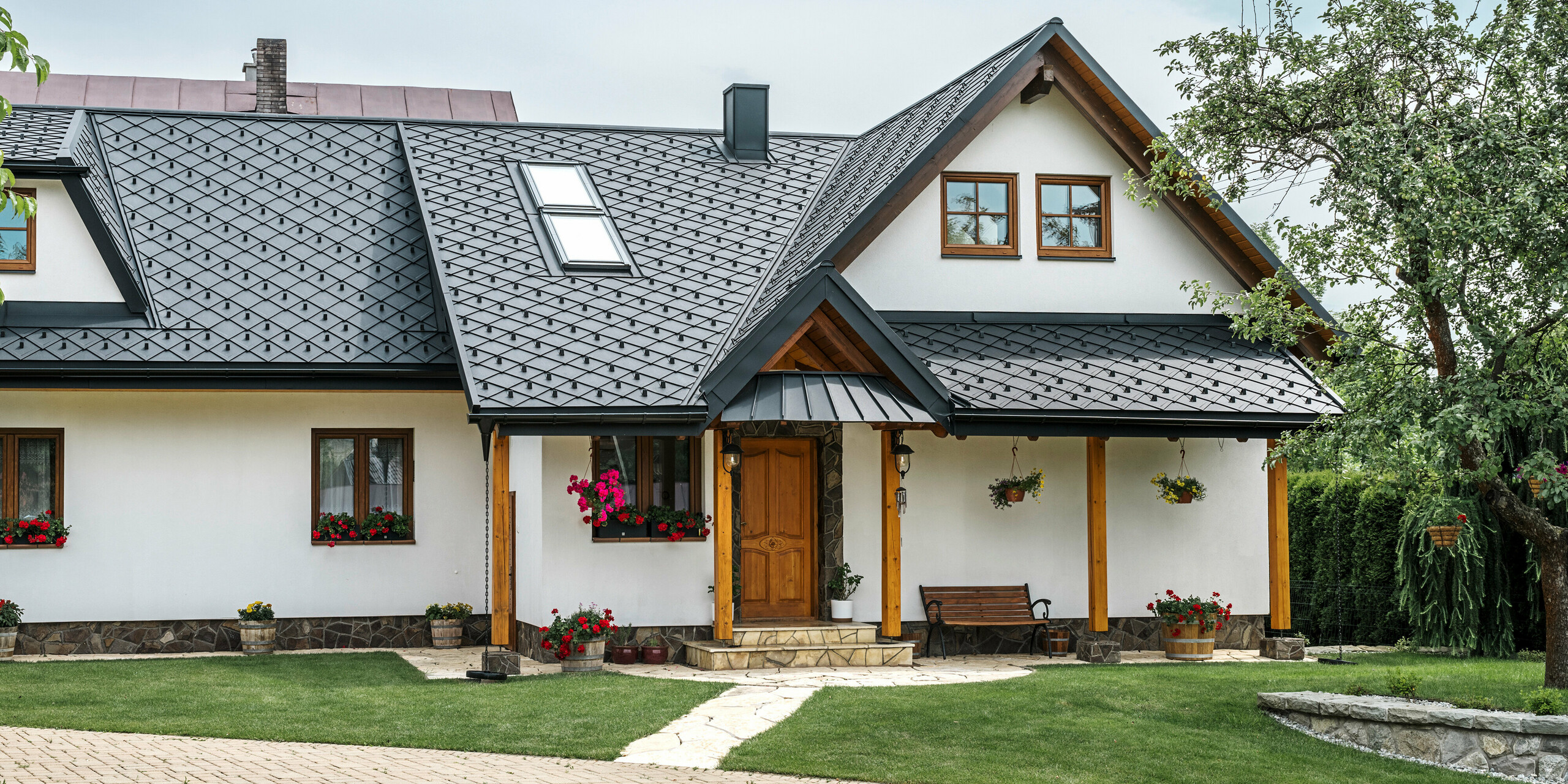 Vstupný priestor tradičného rodinného domu v Starej Bystrici na Slovensku s hliníkovou strechou zo strešných šablón 29 × 29 v čiernej farbe P.10. Súčasťou vysokokvalitnej plechovej strechy s elegantným pultovým vzhľadom sú šikmé strešné okná a veranda s prístreškom oplášteným materiálom PREFALZ. Elegantné a zároveň robustné hliníkové výrobky dodávajú domu moderný a prívetivý vzhľad. Udržiavaná záhrada a kvetinová výzdoba zdôrazňujú, ako dobre je stavba udržiavaná.