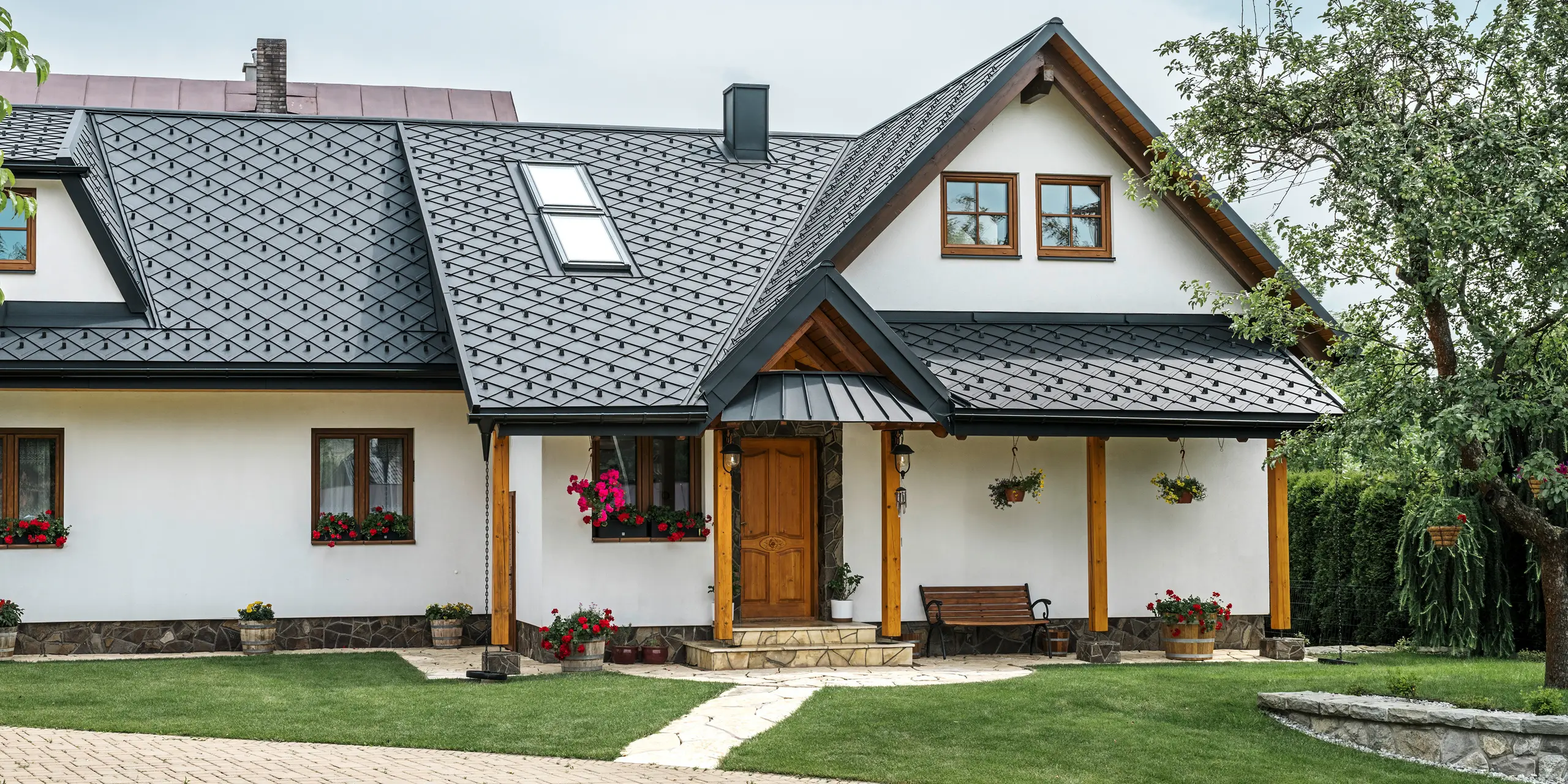 Vstupný priestor tradičného rodinného domu v Starej Bystrici na Slovensku s hliníkovou strechou zo strešných šablón 29 × 29 v čiernej farbe P.10. Súčasťou vysokokvalitnej plechovej strechy s elegantným pultovým vzhľadom sú šikmé strešné okná a veranda s prístreškom oplášteným materiálom PREFALZ. Elegantné a zároveň robustné hliníkové výrobky dodávajú domu moderný a prívetivý vzhľad. Udržiavaná záhrada a kvetinová výzdoba zdôrazňujú, ako dobre je stavba udržiavaná.