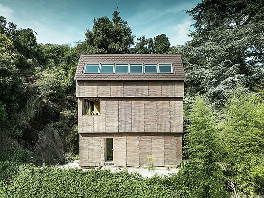 Moderná architektúra vo Francúzsku - dom so strešnými šindľami PREFA v orieškovohnedej farbe P.10 a vŕbovou fasádou, strmo na kopci s veľkorysými strešnými oknami.