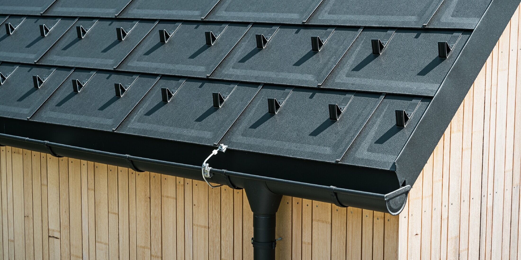 Záber zblízka na PREFA strešný panel R.16 so zachytávačmi snehu, polkruhovým žľabom a dažďovým zvodom v čiernej farbe P.10 vrát. ochrany pred bleskom a zvislej drevenej fasády