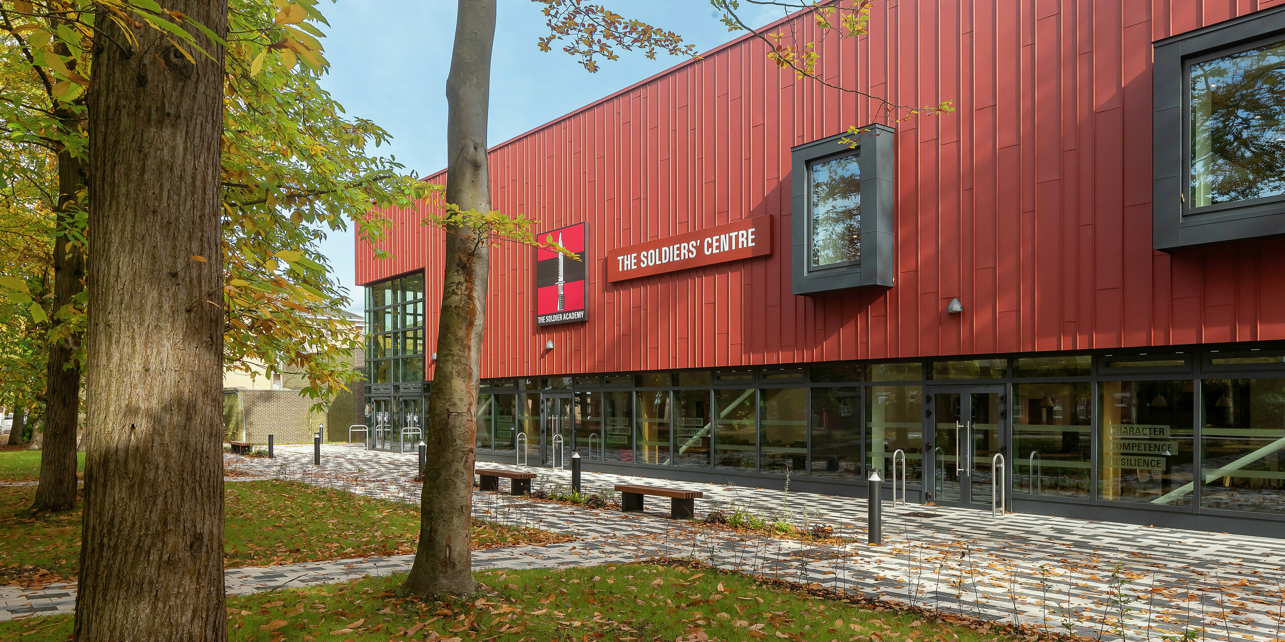 Detailný pohľad na moderné fasádne prvky na Soldiers' Centre v Pirbright: V centre pozornosti je presne položená stojatá falcová obkladová doska z PREFALZu v odtieni P.10 tmavočervená, ktorá v kombinácii s výraznými okennými rámami v P.10 Anthrazit vytvára optický akcent. Jemné línie a hra tieňov podčiarkujú vysokú kvalitu spracovania a inovatívny dizajn hliníkových produktov PREFA – dôkaz udržateľnej a modernej architektúry.