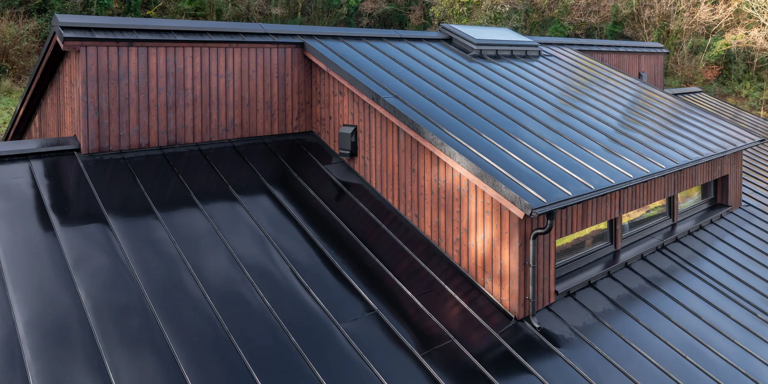 Schrägperspektive auf die Dachlandschaft des House of Holywell in Sligo: Die klar geführten Linien des PREFALZ Aluminiumdachs in P.10 Schwarz verlaufen dynamisch über mehrere Ebenen und lenken den Blick zur präzise ausgeführten Gaube. Die rhythmischen Stehfalze reflektieren das Licht und betonen die hochwertige Verarbeitung sowie die exakten Anschlüsse. In Kombination mit der Holzfassade entsteht ein starkes architektonisches Bild – unterstützt durch die langlebigen, korrosionsbeständigen und wartungsarmen PREFA Aluminiumprodukte.