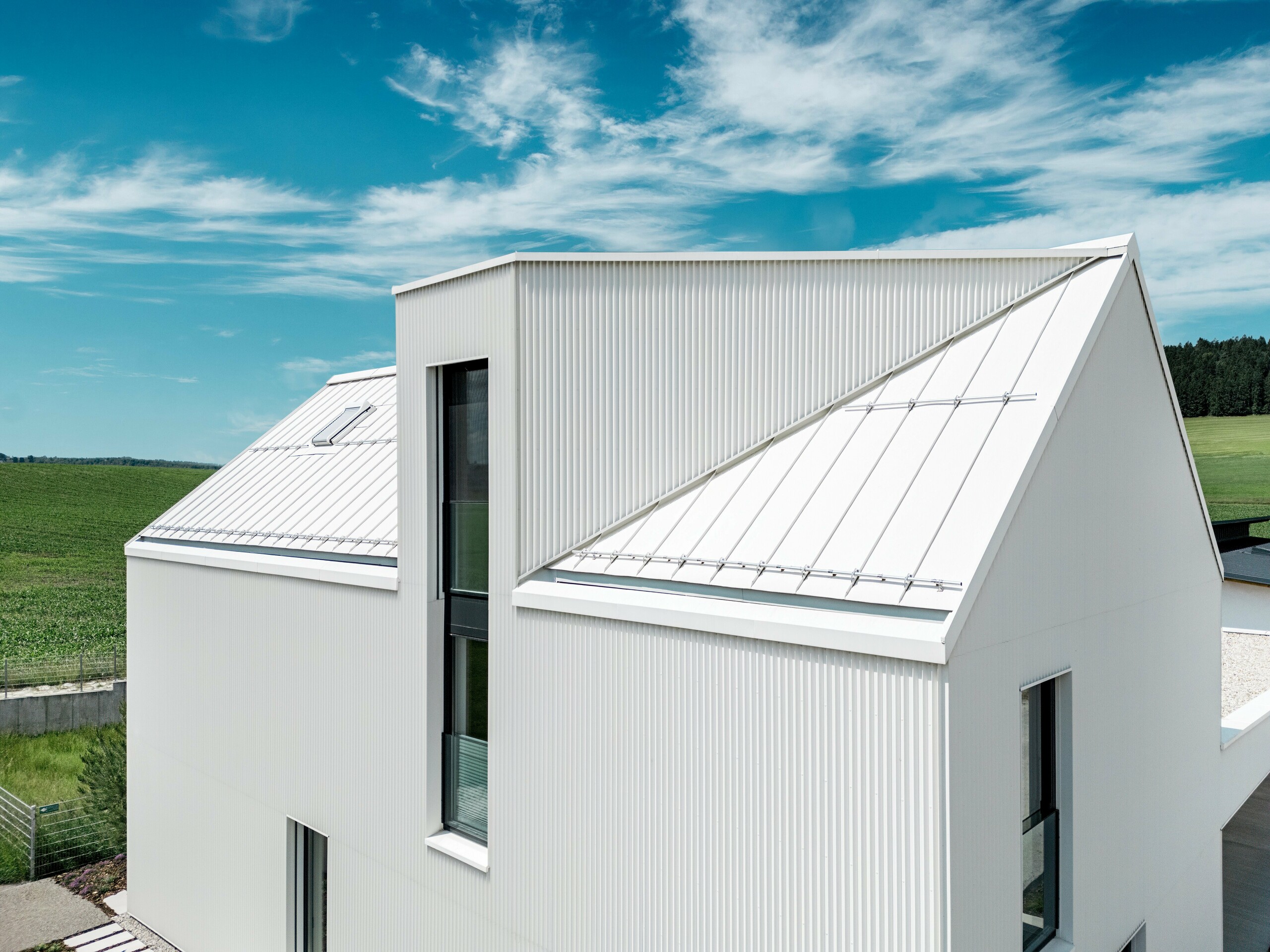 Na obrázku je vidieť strmú, výraznú sedlovú strechu tohto rodinného domu vo Freistadte, ktorá je pokrytá plechom PREFALZ v čisto bielej farbe. Presne zložená fasáda a strecha sú bezšvíkovo spojené, čo vytvára elegantné, čisté línie. Použitý PREFALZ na streche a fasáde zaručuje nielen estetickú stálosť, ale aj vysokú odolnosť voči poveternostným vplyvom. Tým sa budove zaručuje dlhá životnosť a jednoduchá údržba. Súhra moderného dizajnu a funkčných materiálov vytvára harmonickú, modernú architektúru, ktorá je praktická aj vizuálne presvedčivá. Zvlášť zaujímavá je vikierová strecha so šikmým sklonom a oknami siahajúcimi cez dve poschodia.