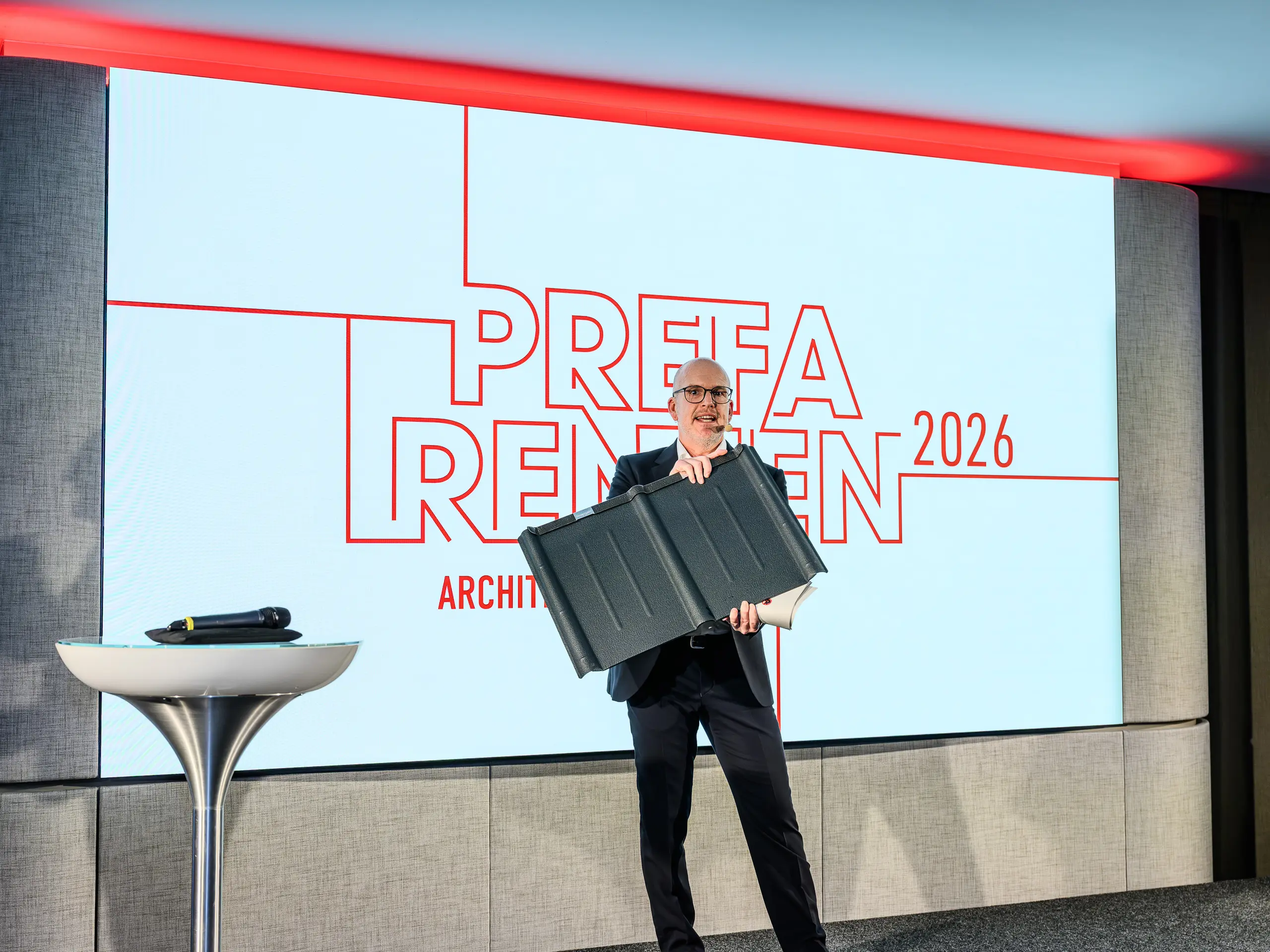 Vrchol prezentácie PREFARENZEN 2026: Jürgen Jungmair, riaditeľ medzinárodného marketingu spoločnosti PREFA, s hrdosťou predstavuje legendárnu strešnú tašku PREFA. V centre pozornosti je tento odolný hliníkový produkt, ktorý sa úspešne vyrába už približne 80 rokov. Moderné pódium s výrazným logom PREFARENZEN podčiarkuje význam tohto produktu pre architektúru orientovanú na budúcnosť. Ľahká, odolná voči búrkam, nerezová – strešná doska PREFA spája techniku a estetiku na najvyššej úrovni.