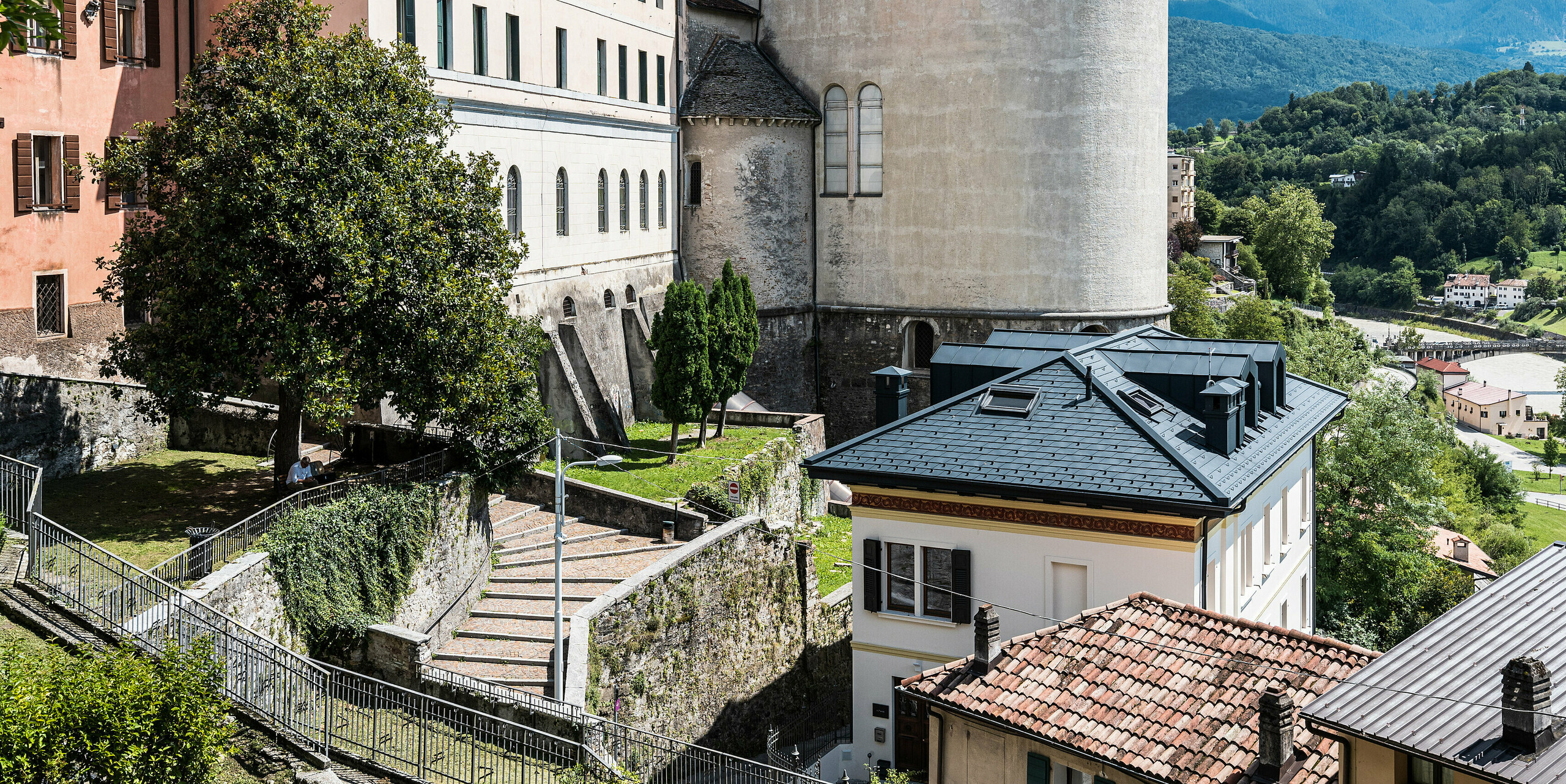 Idylický pohľad na historické centrum mesta Belluno v Taliansku so starostlivo zrekonštruovanou budovou, ktorej strecha je pokrytá hliníkovými šindľami PREFA v antracitovej farbe, ktoré harmonicky ladia s klasickou architektúrou a okolitou zelenou krajinou. Pri rekonštrukcii sa dbá na zachovanie historickej podstaty a zároveň sa integrujú moderné prvky, aby sa dedičstvo zachovalo aj pre budúce generácie.