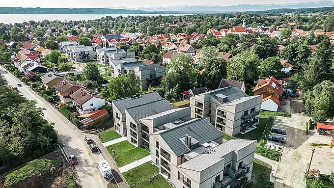 Panoramatický výhľad na veľký pozemok Schmucker v Utting am Ammersee, ktorý sa vyznačuje priestrannou a modernou zástavbou s niekoľkými bytovými domami. Šesť budov je vybavených udržateľnými a odolnými strešnými panelmi PREFA R.16 v tmavosivej farbe a harmonicky zapadajú do okolia. Budovy sú obklopené zelenými stromami a v pozadí sa trbliece modrá farba jazera Ammersee. Architektúra celého rezidenčného projektu zaujme jasnými líniami a estetickou kombináciou drevených fasád a vysokokvalitných hliníkových strešných systémov, ktoré ponúkajú funkčnosť aj vizuálnu eleganciu