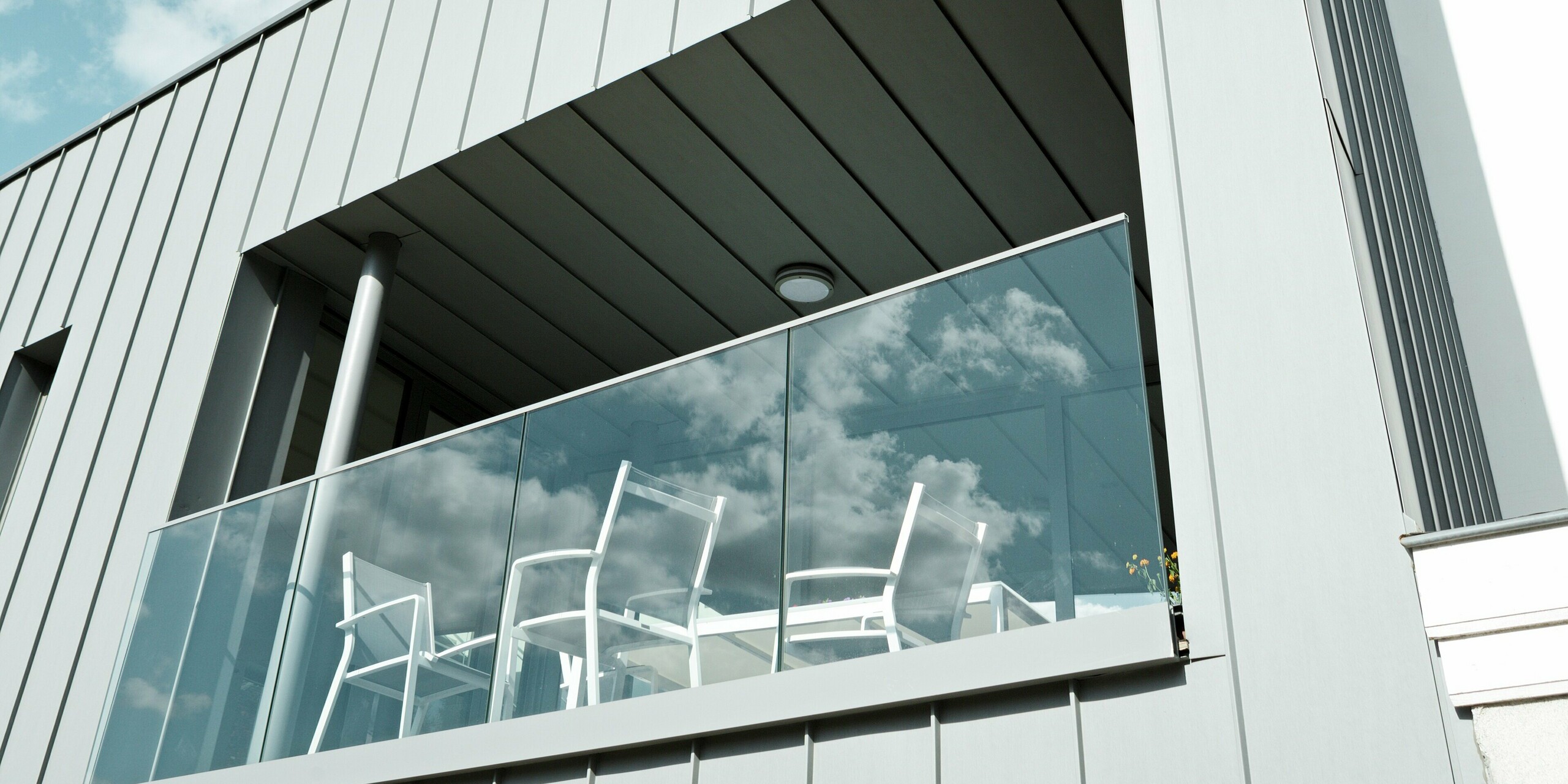 Štýlový detailný záber zastrešenej terasy so skleneným zábradlím na fasáde z PREFALZ v patinovo šedej farbe. Vertikálna konštrukcia zo zliatinového hliníka dodáva budove čisté línie a je navyše odolná voči poveternostným vplyvom, nenáročná na údržbu a trvácna – ideálna pre náročnú architektúru.