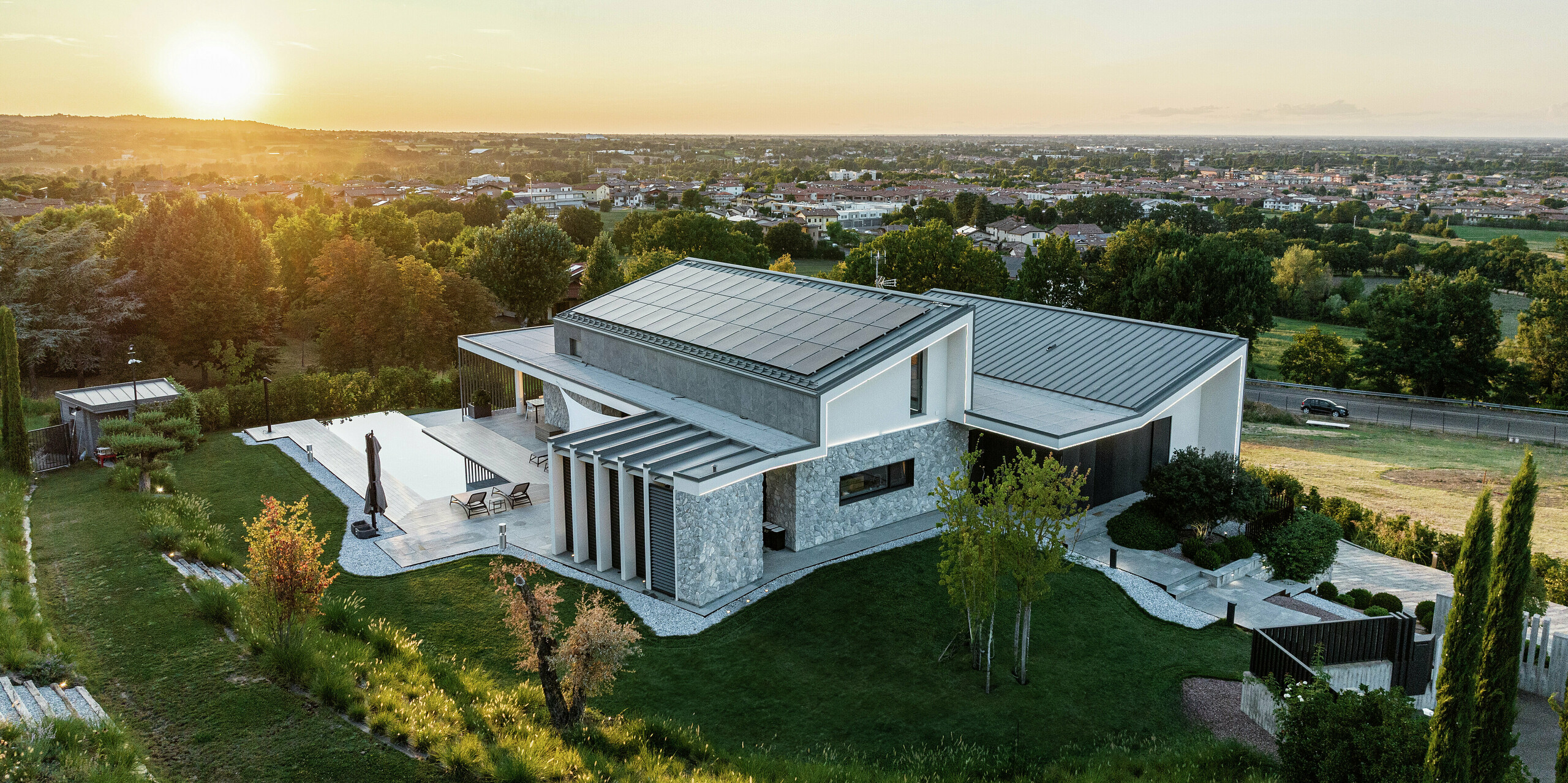 Rodinný dom v Scandiano, Taliansko, v teplom večernom svetle. Dve jednoplášťové strechy z PREFALZu vo farbe P.10 antracit podčiarkujú exkluzívnu architektúru a ponúkajú maximálnu trvanlivosť a odolnosť. Priestranná terasa s bazénom, harmonická záhradná krajina a moderný dizajn fasády dotvárajú celkový luxusný obraz. Veľkoplošný fotovoltický systém zabezpečuje ekologickú výrobu energie zo slnečnej energie.