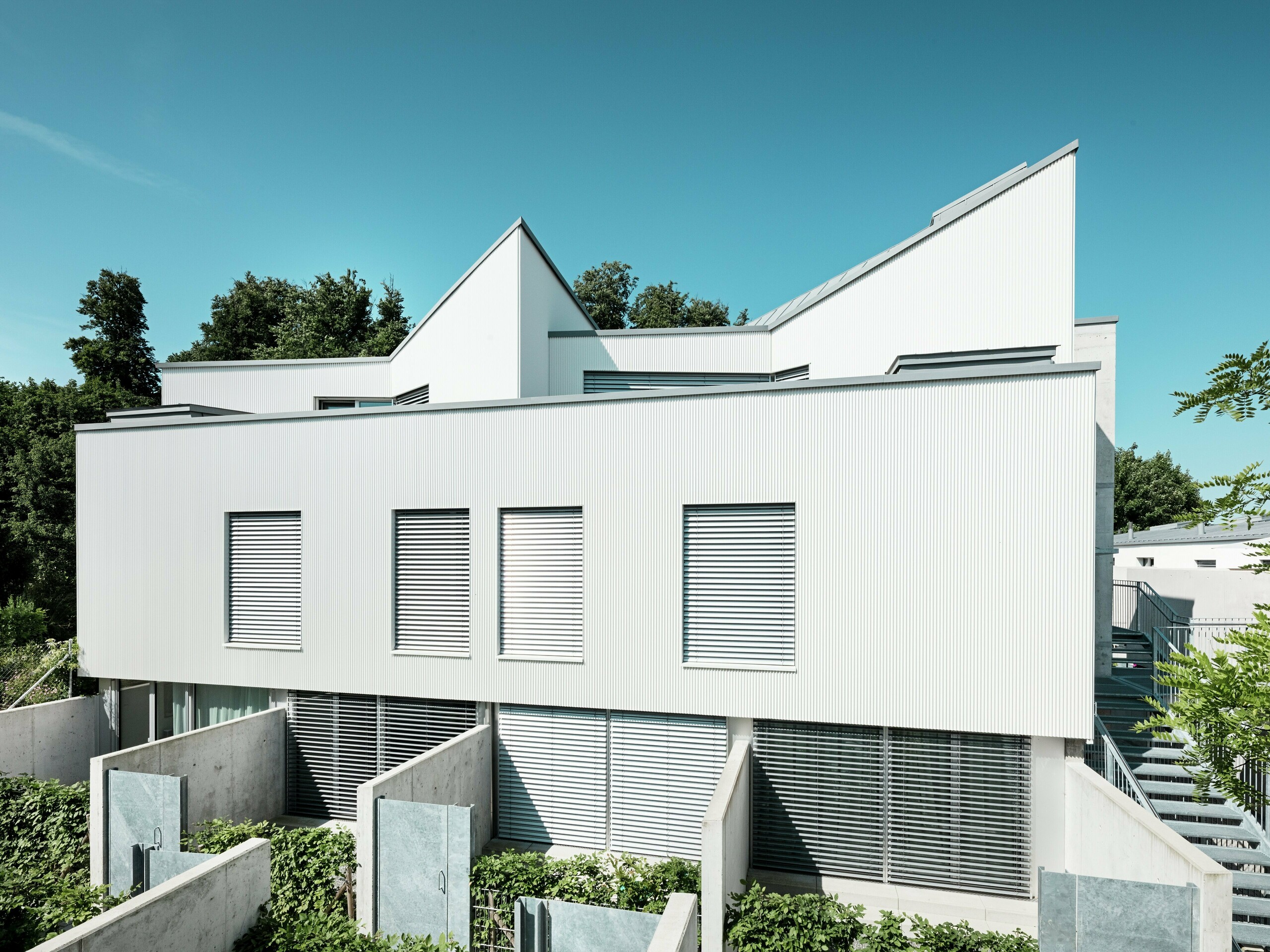 Moderné minidomy v Zollikerbergu vo Švajčiarsku zaujmú svojimi čistými líniami a modernou hliníkovou fasádou PREFA s lisovaným prelamovaným profilom. Vertikálne položené, jemne zakrivené profily vytvárajú výrazný a elegantný vzhľad fasády, ktorý pôsobí živým dojmom vďaka odrazom svetla. Biela povrchová úprava dodáva ľahkosť a zdôrazňuje geometrickú architektúru. Zavesená, odvetraná fasáda trvalo chráni budovu pred vplyvmi počasia a zlepšuje vnútorné prostredie. Je ľahko udržiavateľná a odolná proti korózii.