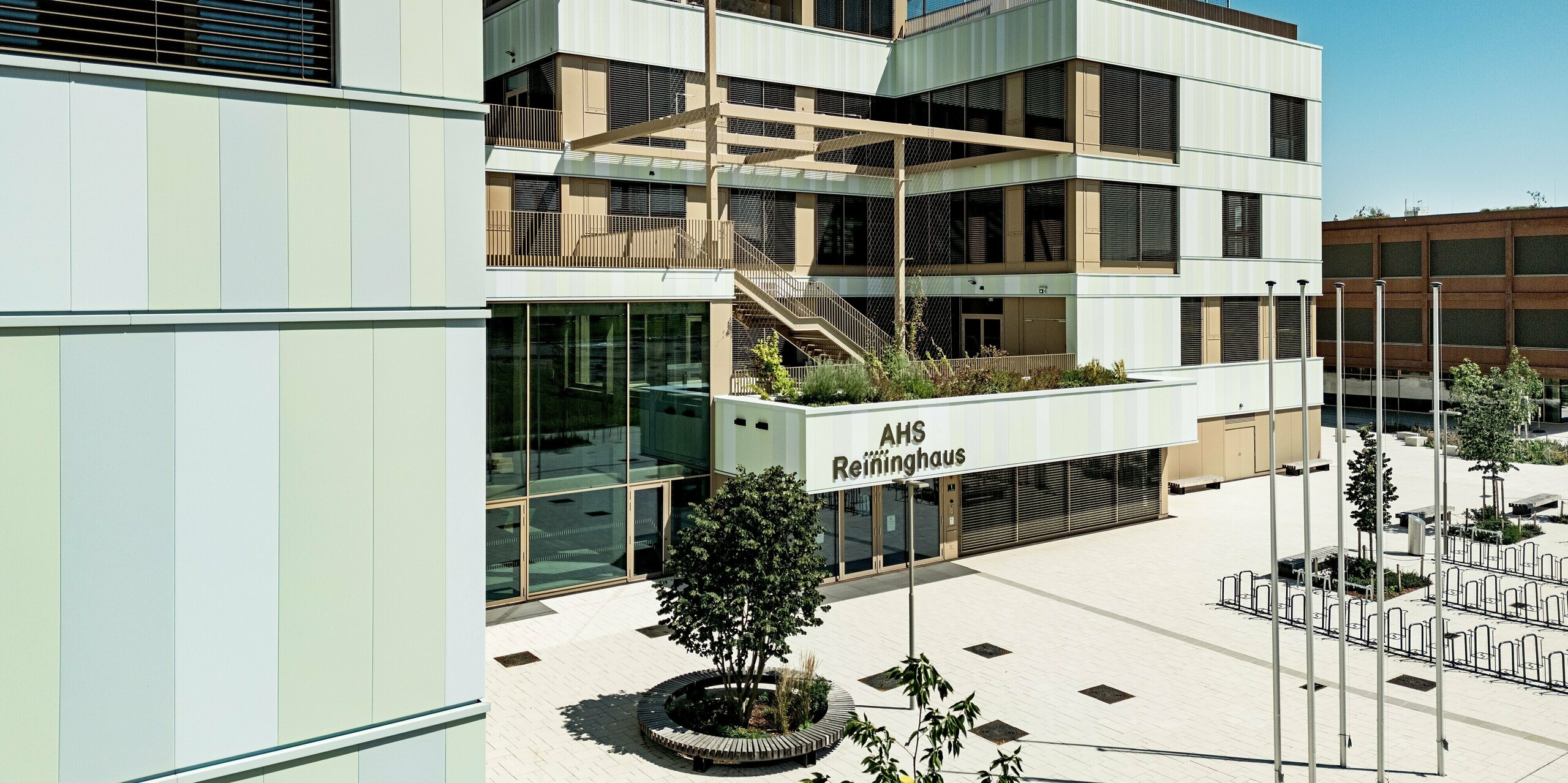 Das Bild zeigt den Eingangsbereich der AHS Reininghaus in Graz. Die Fassade wurde mit langlebigen PREFA-Sidings in Sonderfarben verkleidet. Die vertikal angeordneten Aluminiumprofile in sanften Grüntönen und Weiß sorgen für eine moderne, frische Optik. Die braunen Fensterrahmen und Jalousien ergänzen die Fassade nicht nur optisch, sondern bieten auch funktionalen Sonnenschutz. Die Verwendung von PREFA Sidings macht die Fassade robust, pflegeleicht und wetterbeständig. Sie bietet sowohl ästhetische als auch praktische Vorteile und fügt sich harmonisch in die Umgebung ein.