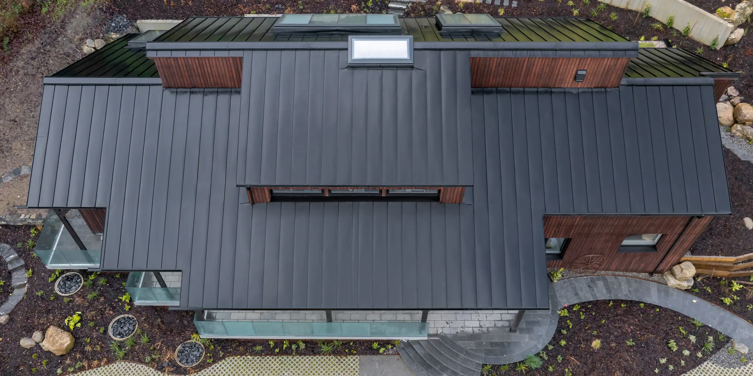 Vogelperspektive auf das House of Holywell in Sligo: Die klar gegliederten Dachflächen des PREFALZ Aluminiumdachs in P.10 Schwarz entfalten aus der Draufsicht ihre volle Wirkung. Die parallel verlaufenden Stehfalze strukturieren das Dach präzise und führen den Blick über Gauben und Ebenen hinweg. Die gleichmäßige Oberfläche betont die hochwertige Ausführung. PREFA Aluminium überzeugt durch Langlebigkeit, Korrosionsbeständigkeit und optimale Funktionalität bei anspruchsvollen Witterungsbedingungen.