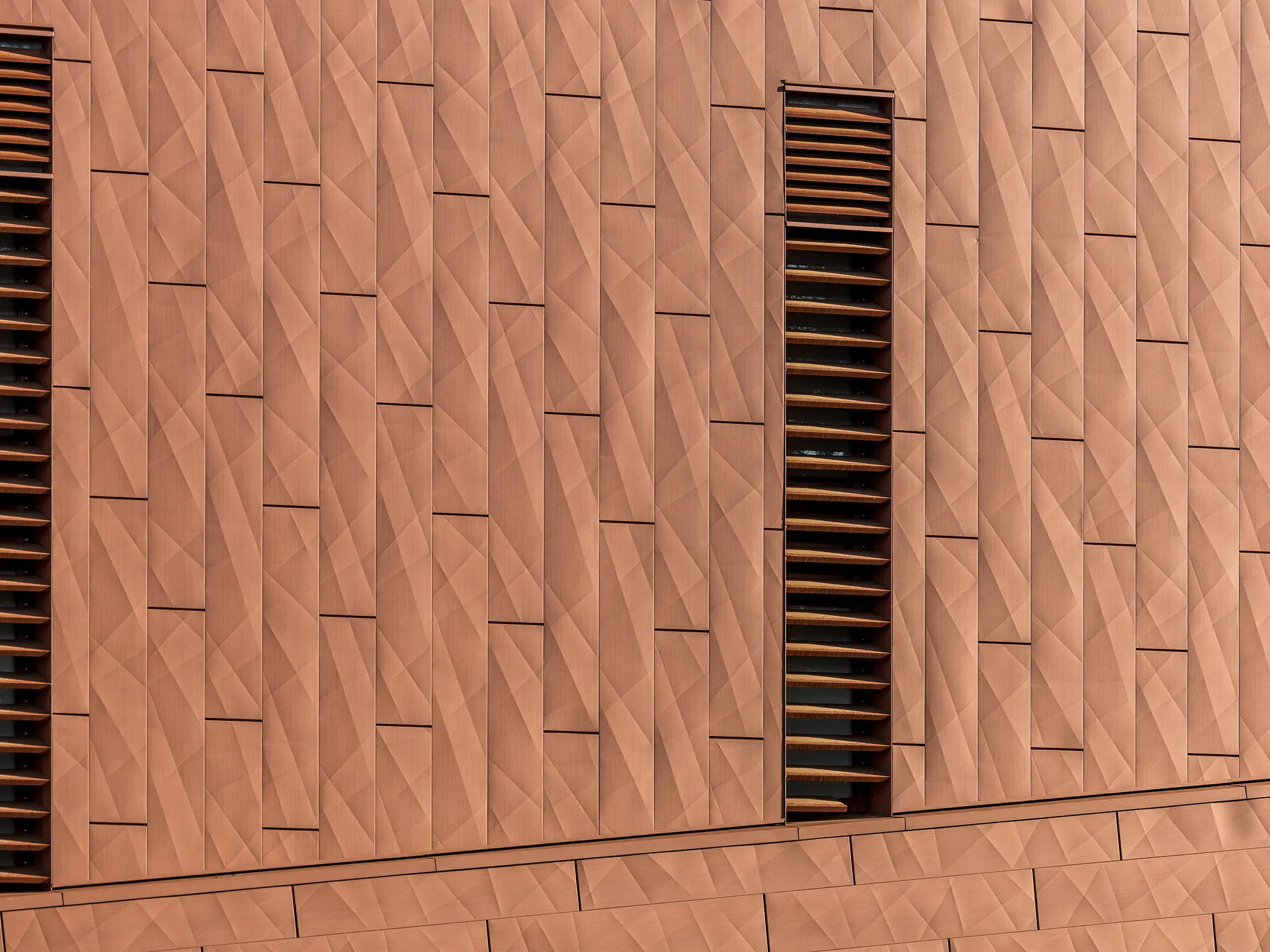 Detailný záber hliníkovej fasády PREFA Siding.X na čerpacej stanici pitnej vody WML v Lottume s jemne zvlneným geometrickým povrchom v teplom vzhľade hrdze. Presne usporiadané panely vytvárajú živú hru svetla a tieňa, zatiaľ čo štíhle vertikálne lamely ako výrazný prvok, ktorý upúta pozornosť, štruktúrujú vzhľad fasády. Ľahké, trvanlivé a úplne recyklovateľné hliníkové panely PREFA presviedčajú vysokou odolnosťou voči poveternostným vplyvom, stálosťou farieb a udržateľným plášťom budovy s nízkymi nárokmi na údržbu.