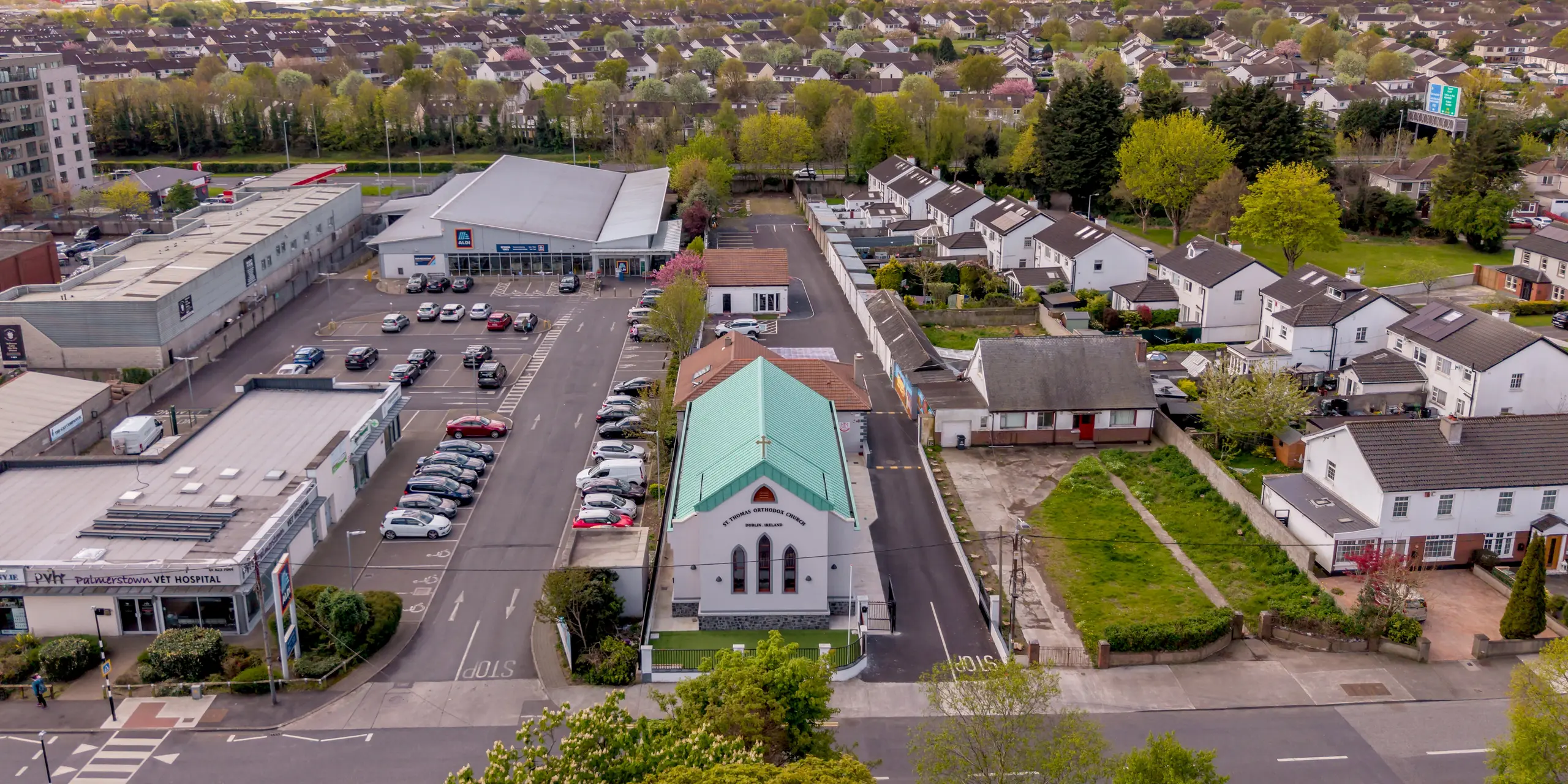 Spektakuläre Vogelperspektive der St. Thomas Orthodox Church in Palmersgate, Dublin, eingebettet in das urbane Umfeld der irischen Hauptstadt. Das weithin sichtbare PREFALZ Aluminium-Stehfalzdach in P.10 Patinagrün bildet einen markanten Blickfang und hebt die Kirche deutlich von den umliegenden Wohn- und Gewerbebauten ab. Die klar gegliederte Dachform, die präzisen Falzlinien und das Kreuz auf dem First verleihen dem Bauwerk eine unverwechselbare Präsenz. Der charakteristische Patinagrün-Ton erinnert an historische Kupferdächer und unterstreicht die sakrale Architektur. Das hochwertige PREFA Aluminium überzeugt dabei durch Langlebigkeit, Korrosionsbeständigkeit, geringes Gewicht und eine nachhaltige, wartungsarme Dachlösung.