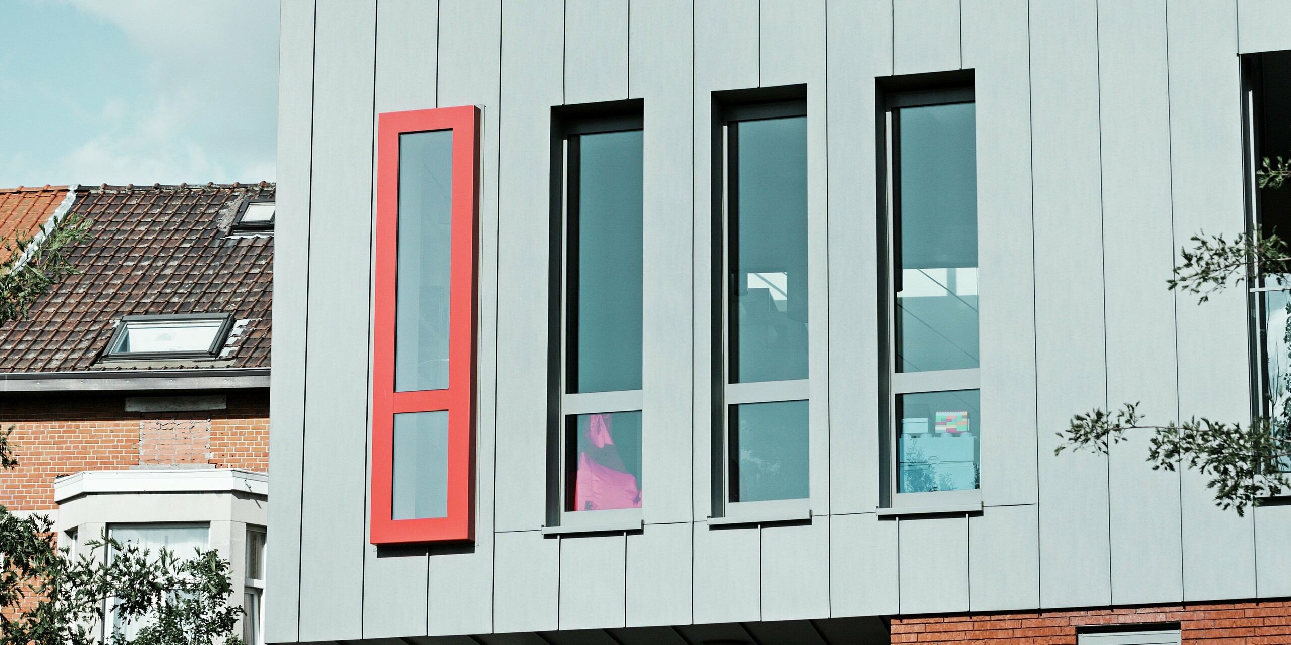 Tento detailný záber ukazuje modernú hliníkovú fasádu PREFALZ v farbe patinovo šedej s vertikálnou stojatou falcovou štruktúrou. Pozornosť upúta najmä úzke okno vo formáte na výšku s výrazným červeným rámom, ktorý dodáva architektúre osobitý akcent. Veľké sklenené plochy zabezpečujú dostatok svetla v budove.