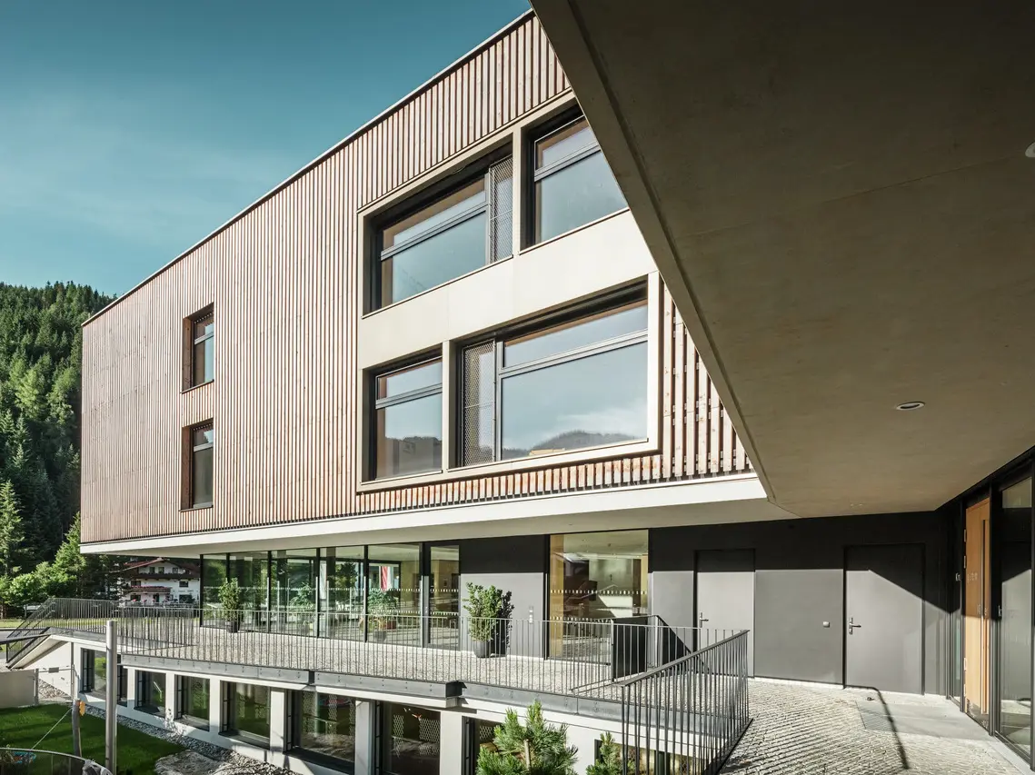 Viacpodlažná budova s moderným architektonickým dizajnom s kombináciou dreva a omietky na fasáde v alpskom prostredí so zalesnenými kopcami v pozadí. Budova má veľké okná, ktoré prepúšťajú veľa svetla, a krytú terasu, ktorá zvýrazňuje vstup.