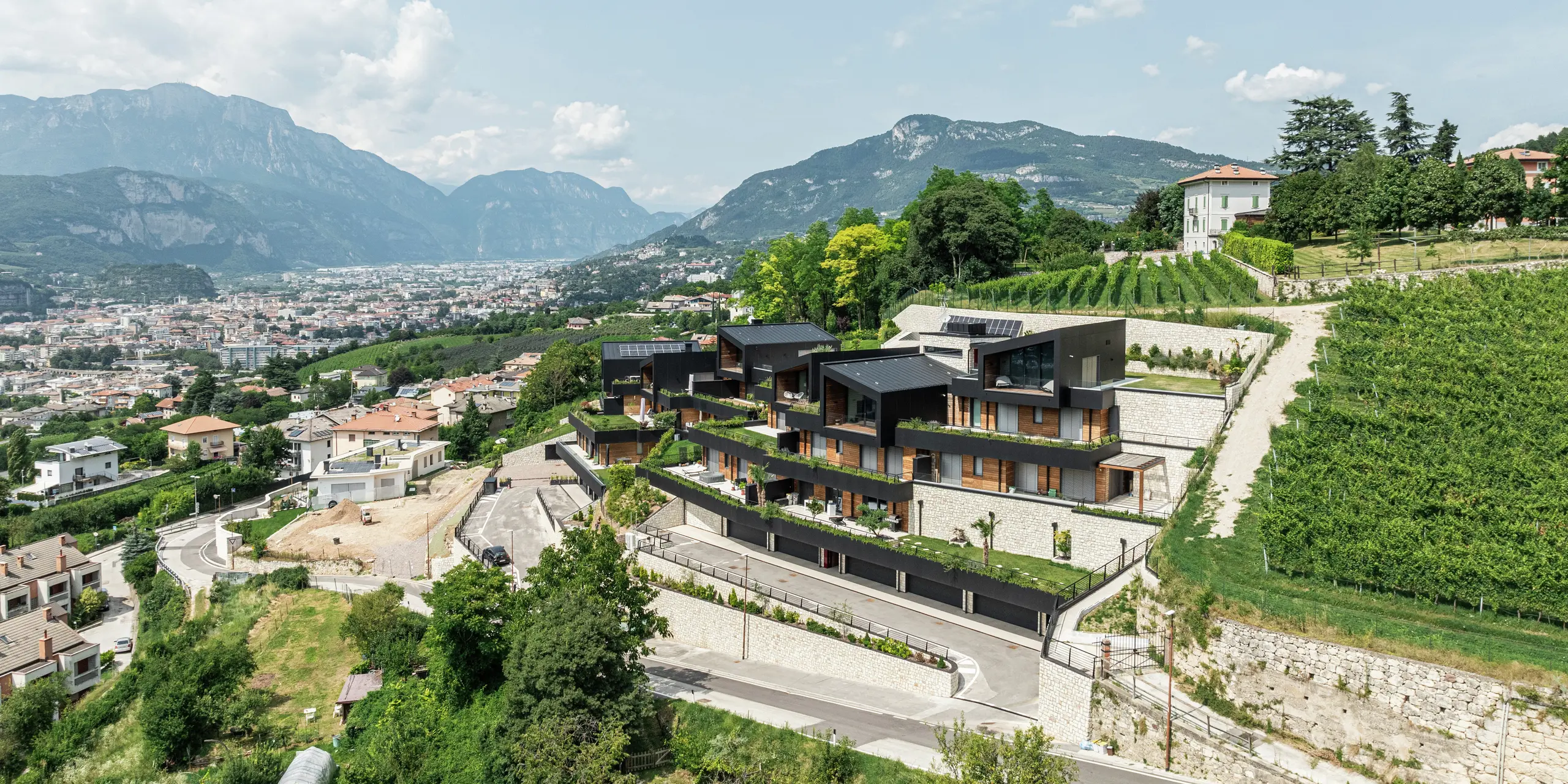 Nádherná poloha na svahu s výhľadom na Trento: exkluzívny rezidenčný komplex dokonale zapadá do okolitého vinohradníckeho a kopcovitého krajiny. Moderná architektúra spája prírodný kameň, drevo a odolnú hliníkovú fasádu PREFA v čiernej farbe P.10 do elegantného a odolného celku. Strecha PREFALZ v čiernej farbe P.10 zaisťuje maximálnu ochranu a dlhú životnosť. Rozľahlé terasy a zeleň podčiarkujú udržateľný koncept bývania a vytvárajú harmonické spojenie prírody a modernej architektúry.