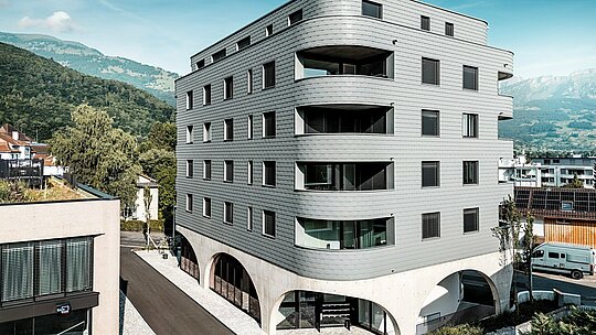 Vonkajší pohľad na Bernerhaus v Buchsi s výrazným trojuholníkovým tvarom a jemne zaoblenými balkónovými pásmi. Fasáda z PREFA fasádnych šindľov v odtieni P.10 svetlošedá obklopuje budovu ako šupinatá hliníková koža a vytvára presný, živý obraz fasády. Nad masívnym betónovým podstavcom s elegantnými arkádami pôsobí ľahká, odolná proti poveternostným vplyvom hliníková fasáda PREFA obzvlášť výrazne. Trvanlivá, nenáročná na údržbu a tvarovo stabilná, vytvára moderný architektonický dojem na pozadí alpských vrcholov.