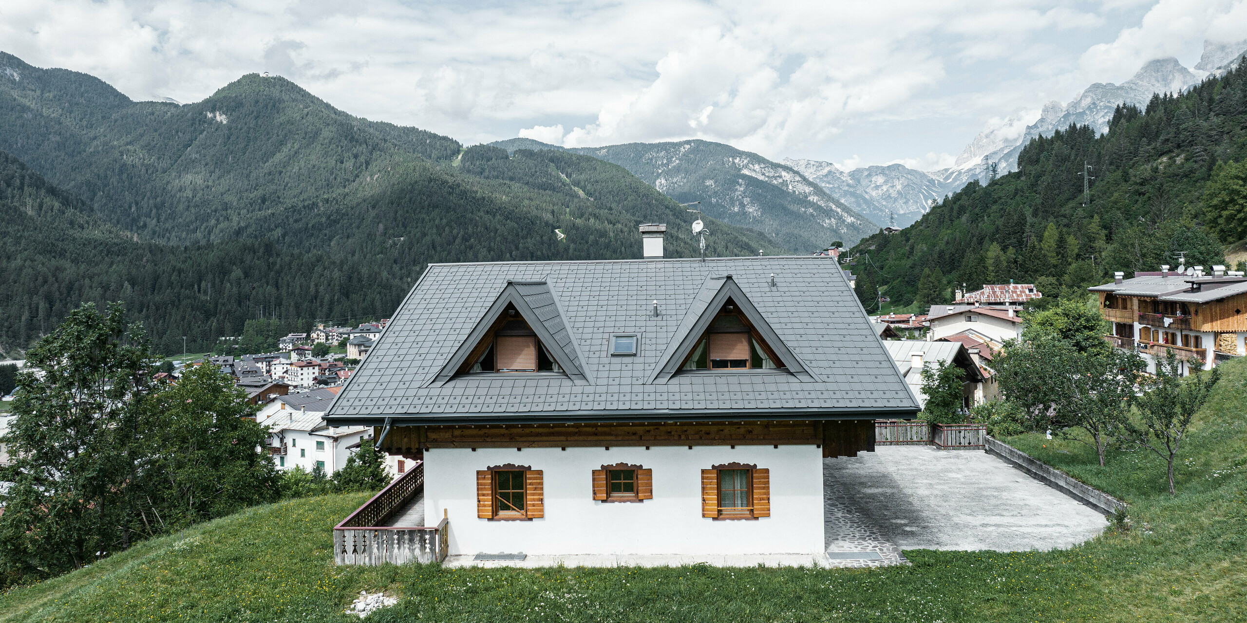 Na zadnej strane horskej chaty v Auronzo di Cadore v Taliansku je čerstvo položená falcovaná škridla PREFA vo svetlošedej farbe. Dom stojí na zelenej lúke s majestátnymi Dolomitmi v pozadí. Dva špicaté vikiere dokonale splývajú s horskou kulisou v pozadí. Starostlivá obnova strechy zvýrazňuje tradičný šarm budovy, zatiaľ čo moderné prvky ju spájajú s alpskou krajinou.