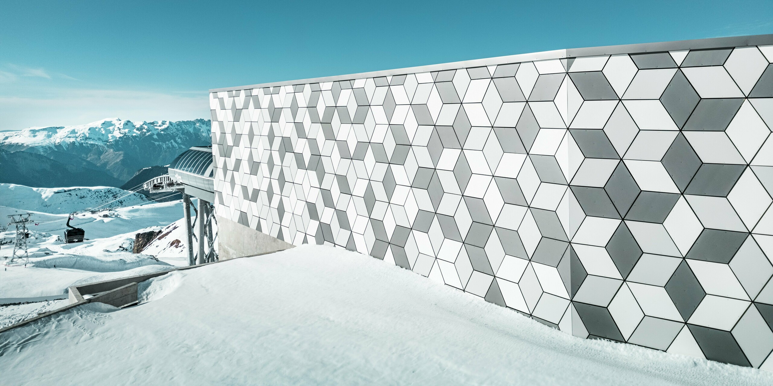  Fotografia hliníkovej fasády stanice lanovky v Les Deux Alpes: Výrazná fasáda z hliníkových kompozitných panelov PREFABOND zaujme svojím pútavým geometrickým usporiadaním kosoštvorcov. Harmonická kombinácia bielej a sivej farby vytvára moderný vzhľad, ktorý dokonale zapadá do majestátnej horskej krajiny. Za fasádou je vidieť žiarivo modrú oblohu a majestátnu horskú panorámu.