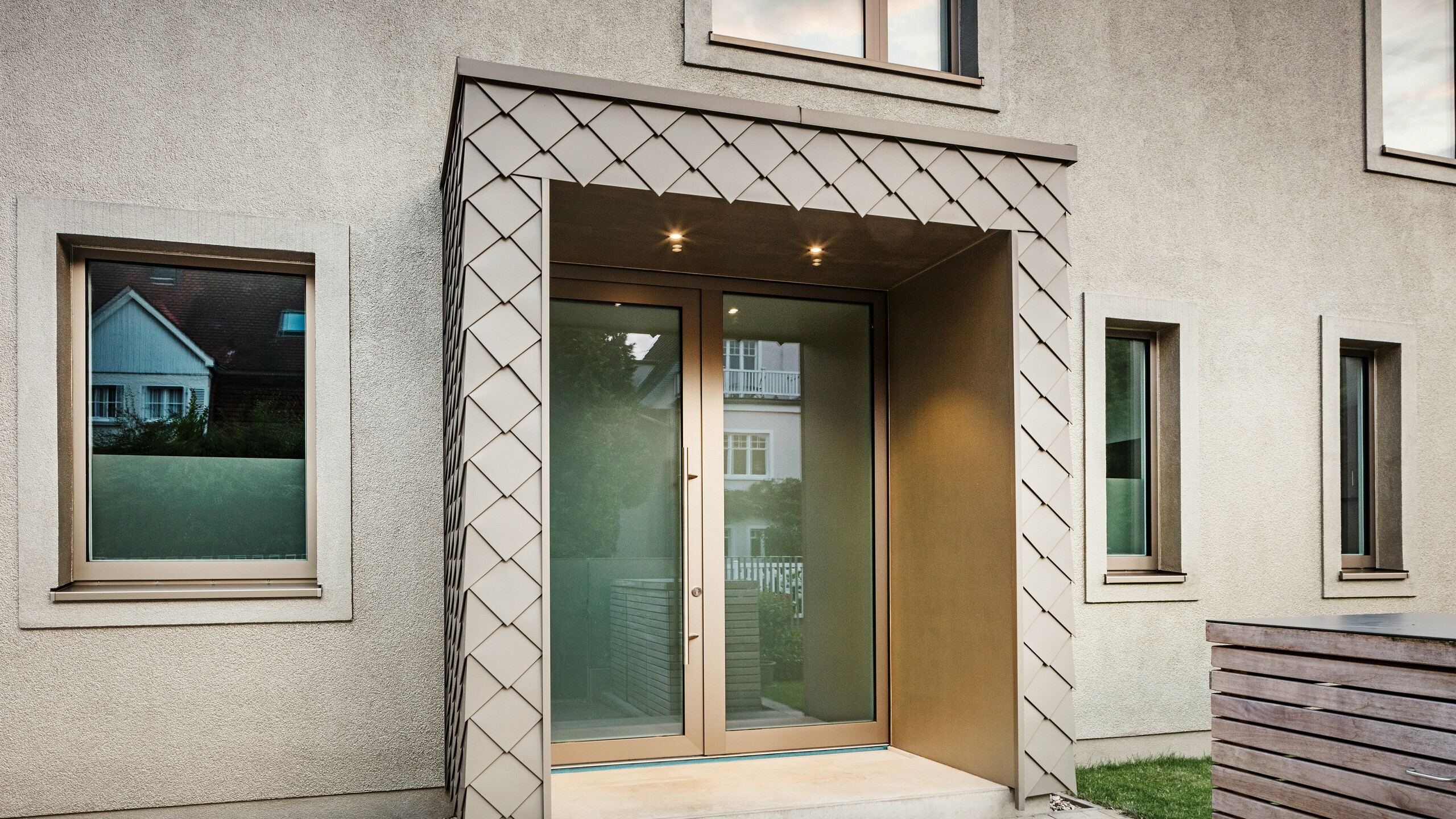 Detailný záber na vstupný priestor moderného rodinného domu s hliníkovým obkladom bronzovej farby od spoločnosti PREFA. Vchod je zakrytý a obložený elegantnými hliníkovými šablónami, ktoré dodávajú priestoru moderný a štýlový vzhľad. Naľavo a napravo od vstupných dverí sú veľké okná, ktoré vytvárajú svetlú a príjemnú atmosféru.