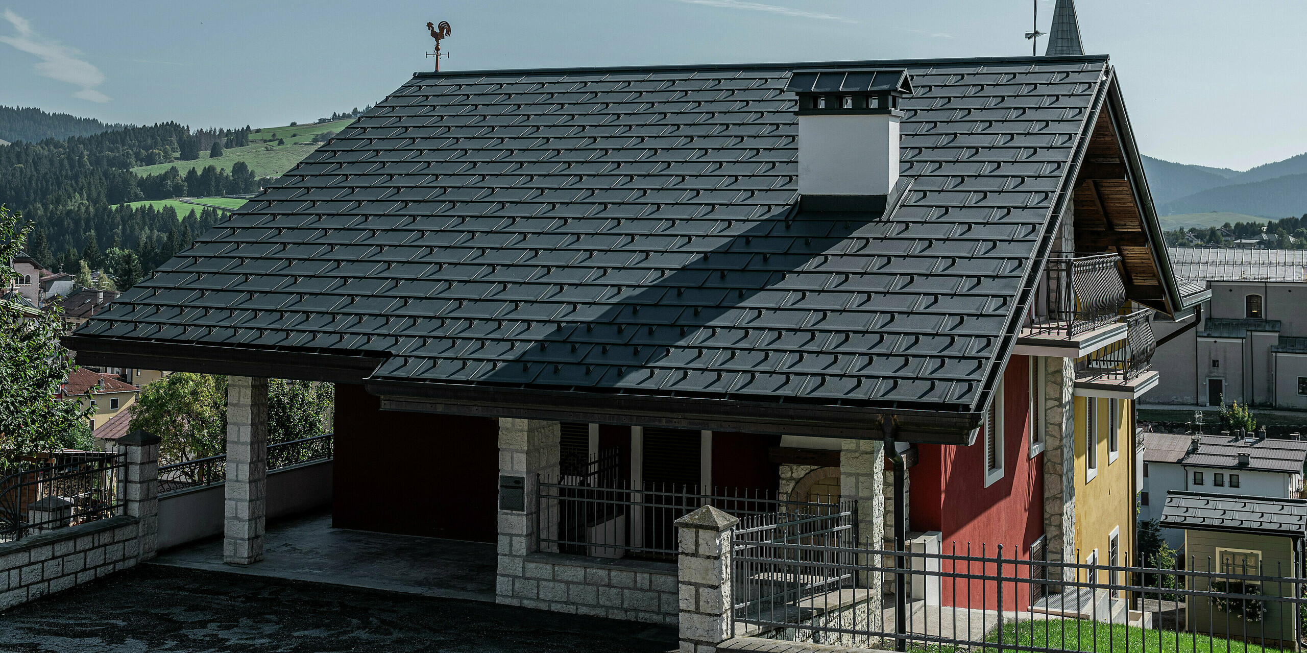 Detailný pohľad na vstupnú časť rodinného domu v Gallio s robustnou strechou PREFA zo strešných panelov R.16 v tmavošedej farbe P.10. Strešná krytina z hliníka zaisťuje elegantný vzhľad a zároveň ponúka najvyššiu ochranu pred poveternostnými vplyvmi. Presné spracovanie a odolný materiál zaručujú bezúdržbové riešenie na desiatky rokov. Masívny komín s obkladom z prírodného kameňa a tradičná veterná ružica dodávajú štýlový akcent. Perfektná kombinácia modernej techniky a alpského šarmu.