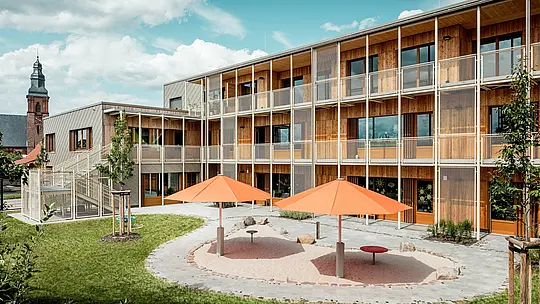 Zadná časť materskej školy v Alzenau zobrazuje chránený vnútorný dvor s nadväzujúcim herným areálom. Dva oranžové slnečníky vytvárajú farebné akcenty nad kruhovým pieskoviskom. V pozadí sa rozprestiera obytný trakt realizovaný v udržateľnej masívnej drevostavbe s predsadenou, filigránovou balkónovou a schodiskovou konštrukciou z kovu. Na ľavej strane je viditeľná hliníková fasáda s bronzovými PREFA fasádnymi šablónami 44 × 44, ktoré poskytujú odolnú a esteticky hodnotnú ochranu proti poveternostným vplyvom. Kombinácia hliníka, dreva a ocele je symbolom inteligentného prepojenia funkčnosti, udržateľnosti a architektonickej kvality. Pohľad na kostol v pozadí urbanisticky zakotvuje projekt v historicky formovanom obraze obce Alzenau.