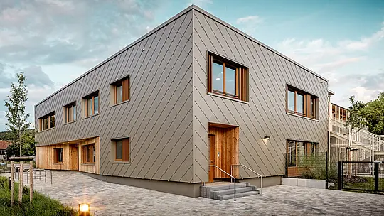 Moderná materská škola v Alzenau zaujme svojou čistou architektúrou a kvalitnou hliníkovou fasádou PREFA v bronzovej farbe. Fasáda z hliníkových šablón 44 × 44 dodáva budove elegantný, štruktúrovaný vzhľad a vizuálne spája jednotlivé funkčné časti do harmonickej jednoty. Drevené obklady pri vstupoch vytvárajú teplý, prirodzený kontrast k trvácnemu hliníku. Kombinácia masívnej stavby, drevených prvkov a zavesenej prevetrávanej fasády je príkladom udržateľného stavebného riešenia. Nenáročný hliníkový plášť PREFA presviedča nielen svojou recyklovateľnosťou, ale aj odolnosťou voči poveternostným vplyvom – ideálny pre perspektívne vzdelávacie stavby s vysokými architektonickými nárokmi.