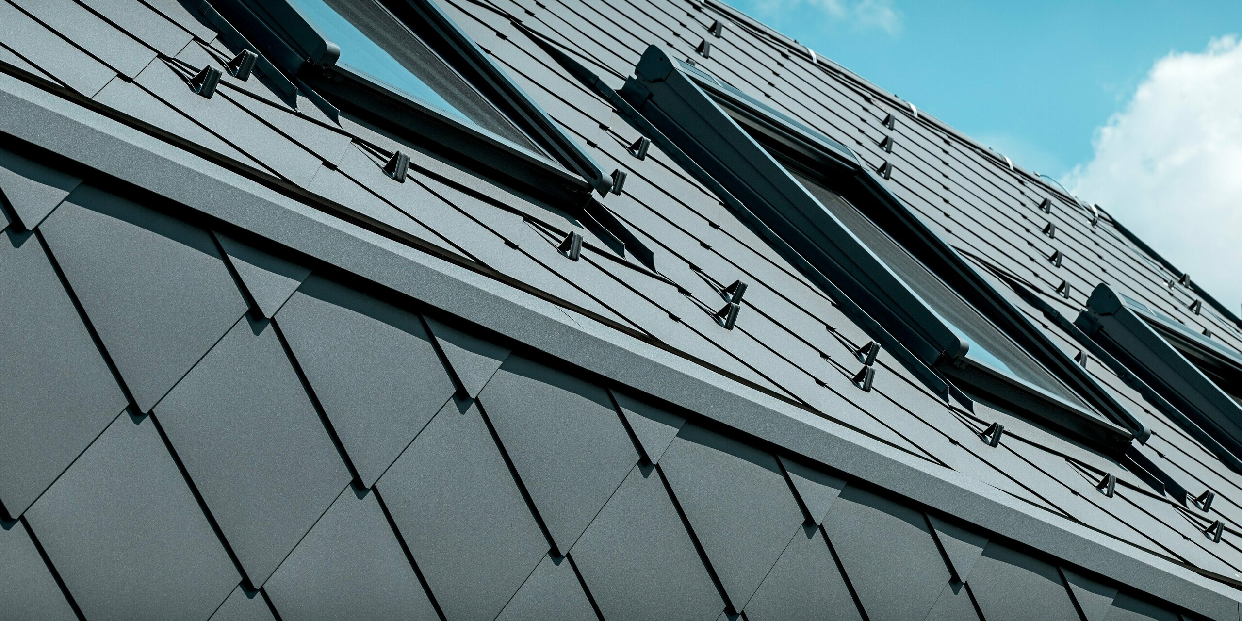 Na obrázku je dynamický detail strechy rodinného domu v Trnave, pokrytej strešnými šablónami PREFA 44 × 44 v odtieni P.10 tmavošedá. Niekoľko strešných okien dodáva streche moderný akcent a elegantným spôsobom narúša jednotnú štruktúru hliníka. Čistá pokládka, integrované snehové zábrany a odolný povrch podčiarkujú vysokú kvalitu produktov PREFA. Trvanlivá hliníková krytina ponúka optimálnu ochranu pred poveternostnými vplyvmi, tvarovú stabilitu a výrazný, moderný vzhľad v kontraste s jasnou oblohou.