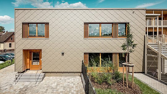 Zadná strana modernej materskej školy v Alzenau je zobrazená z čelnej perspektívy a zaujme čistými líniami a výraznou fasádou z bronzovo sfarbených PREFA fasádnych šablón 44 × 44. Geometrická štruktúra fasády vytvára harmonický šupinový vzor, ktorý je akcentovaný pokojnou horizontálnou orientáciou okien. Veľkorysé drevené rámy a vertikálne lamely dodávajú prírodné akcenty a vytvárajú teplú, prívetivú atmosféru. Premyslená kombinácia trvácneho hliníka a regionálneho dreva predstavuje udržateľnú architektúru s vysokou estetickou kvalitou. Snímka zdôrazňuje precízne vyhotovenie, kvalitný výber materiálov a úspešné začlenenie budovy do mestského prostredia.