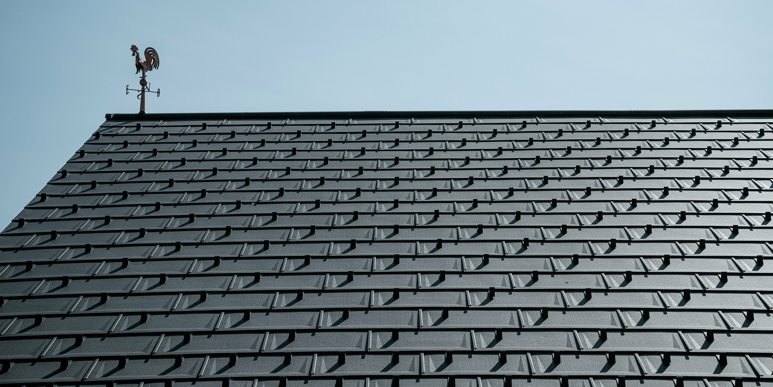 Detailný pohľad na strechu PREFA s strešnými panelmi R.16 v odtieni P.10 tmavošedá. Čistý, moderný vzhľad hliníkových strešných panelov zaručuje trvácne, odolné voči poveternostným vplyvom a bezúdržbové strešné riešenie. Snehové zábrany poskytujú dodatočnú bezpečnosť v regiónoch s veľkým množstvom snehu. Strechu korunuje klasický veterný ukazovateľ, ktorý spája tradíciu s modernou technikou. Vďaka robustným strešným panelom PREFA R.16 zostáva strecha stabilná a estetická po celé desaťročia.