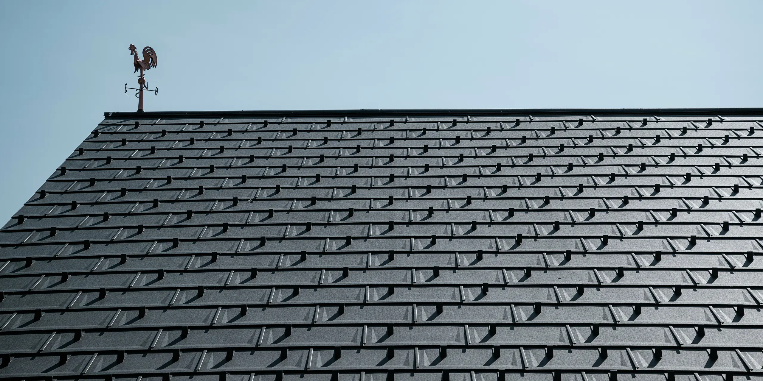 Detailný pohľad na strechu PREFA s strešnými panelmi R.16 v odtieni P.10 tmavošedá. Čistý, moderný vzhľad hliníkových strešných panelov zaručuje trvácne, odolné voči poveternostným vplyvom a bezúdržbové strešné riešenie. Snehové zábrany poskytujú dodatočnú bezpečnosť v regiónoch s veľkým množstvom snehu. Strechu korunuje klasický veterný ukazovateľ, ktorý spája tradíciu s modernou technikou. Vďaka robustným strešným panelom PREFA R.16 zostáva strecha stabilná a estetická po celé desaťročia.