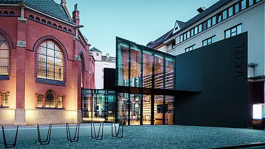 Obrázok ukazuje impozantnú architektúru v Olomouci, ktorá spája historický Červený kostol s modernou novostavbou. Výrazná čierna fasáda z hliníkových kompozitných dosiek PREFABOND na novostavbe dokonale kontrastuje s červenými tehlovými múrmi kostola. Veľké sklenené plochy umožňujú nahliadnuť do interiéru novostavby, kde sú viditeľné police s knihami a príjemná atmosféra. Presné spracovanie materiálov vytvára harmonickú rovnováhu medzi historickými a modernými prvkami. Použitie odolných a esteticky príťažlivých materiálov, ako je PREFABOND, zaisťuje nielen moderný vzhľad, ale aj trvalú kvalitu a funkčnosť. Táto architektúra pôsobivým spôsobom ukazuje, ako sa staré a nové môžu spojiť a vytvoriť jedinečný mestský celok.