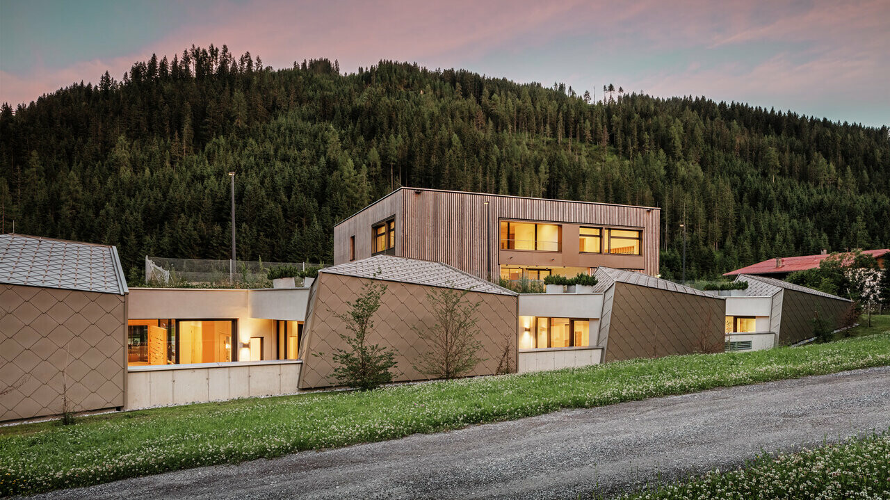 Na obrázku je vzdelávacie centrum Gerlos za súmraku s vnútorným osvetlením. Budova s moderným dreveným obložením a hliníkovými šablónami harmonicky zapadá do prírodného prostredia hustého ihličnatého lesa v pozadí. Obloha sa kúpe v jemných večerných tónoch a vytvára pokojnú a prívetivú atmosféru.