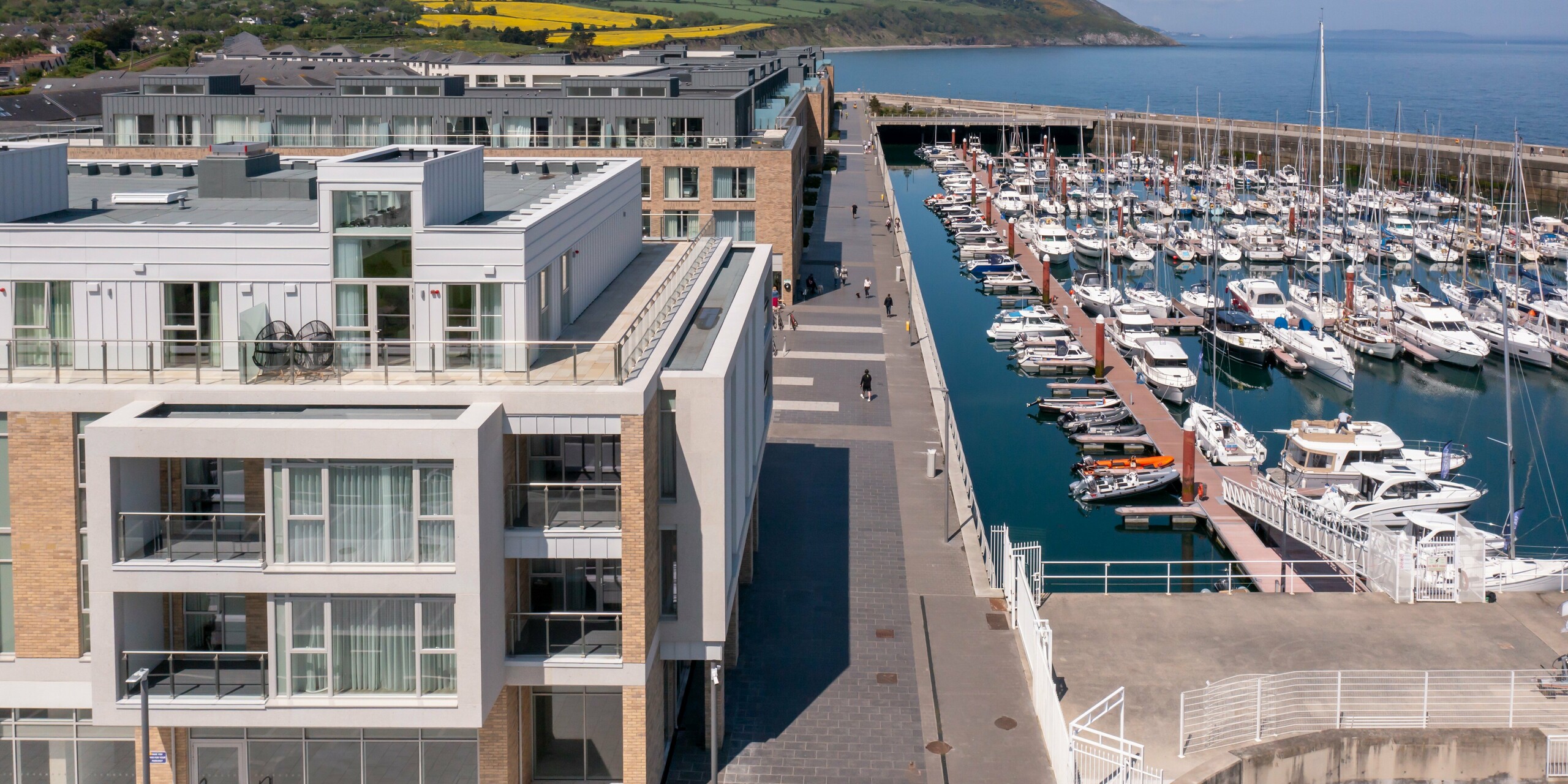 Letecký pohľad na Marina Village Greystones vo Wicklow, Írsko - moderná rezidenčná výstavba priamo pri prístave. Fasáda obytného komplexu je navrhnutá z približne 2 000 m² robustného materiálu PREFALZ vo farbách P.10 prefa biela a P.10 svetlošedá. Elegantná architektúra s veľkorysými presklenými priečeliami a balkónmi, obklopená malebnou krajinou pri vode. Vysokokvalitný hliníkový obklad PREFA ponúka odolnú ochranu a nadčasovú estetiku.