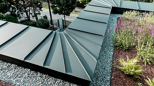 Záber zachytáva detailný pohľad na riešenie atiky budapeštianskej viacgeneračnej vily, obloženej hliníkom PREFALZ v odtieni P.10 antracit. Vlnovito (vejárovo) usporiadané pásy sa presne spájajú na hrane budovy a demonštrujú vysokú tvarovateľnosť a kvalitu spracovania hliníkových produktov PREFA. Antracitový kov vytvára elegantný kontrast k zelenému strešnému priestoru s kamenným štrkom a kvitnúcimi rastlinami. Scéna zdôrazňuje dlhú životnosť, odolnosť voči poveternostným vplyvom a technickú prepracovanosť riešenia PREFA.