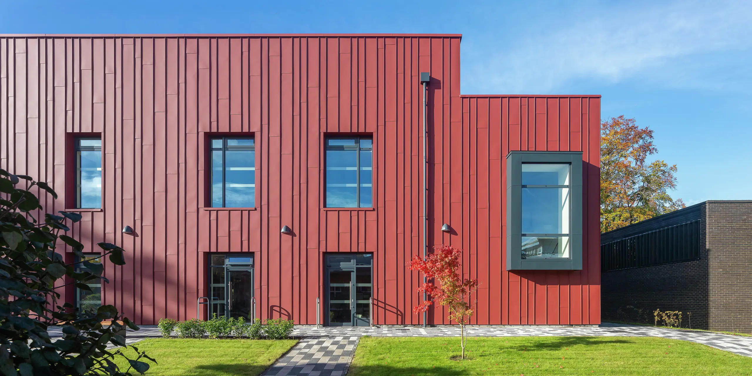 Moderná architektúra s PREFALZ: Fasáda Soldiers' Centre v Pirbrightu zaujme elegantným obložením z PREFALZ v odtieni P.10 tmavočervená. Štýlové okenné rámy v odtieni P.10 Anthrazit dodávajú budove dynamický vzhľad. Trvanlivé, estetické a udržateľné – hliníkové produkty PREFA pre inovatívne stavebné koncepty.