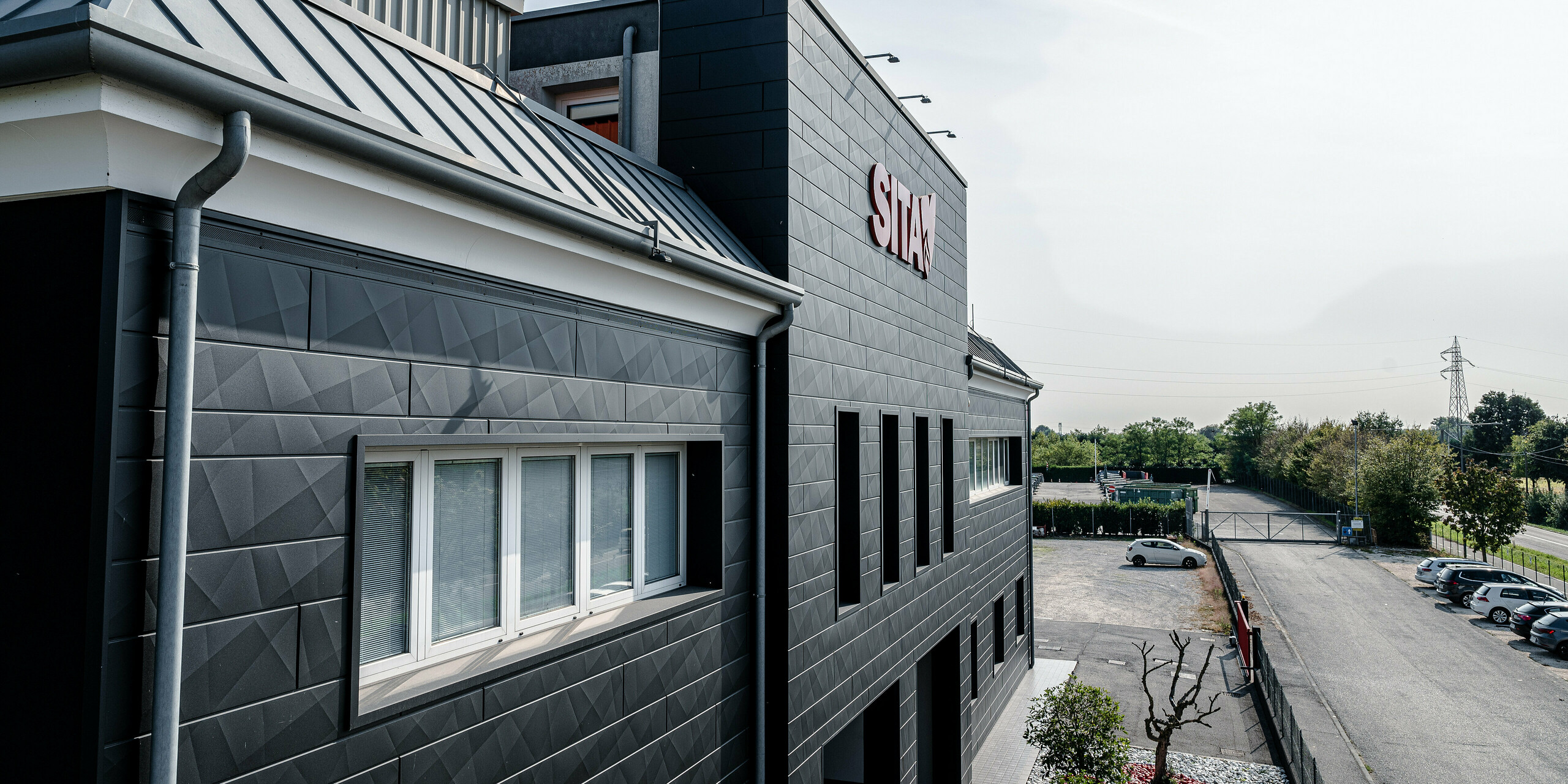 Perspektívny pohľad na budovu spoločnosti SITA v Porcellengu v Taliansku so zameraním na štruktúrovanú fasádu PREFA Siding.X v čiernej farbe P.10. Kompozícia snímky ukazuje harmonické začlenenie budovy do okolia s jasnými líniami a výraznou geometriou strechy, ktorá odráža vysokú kvalitu a povedomie o dizajne spoločnosti PREFA. Červené logo spoločnosti tvorí na fasáde pútavý akcent, ktorý je nielen vizuálne príťažlivý, ale aj funkčný a odolný voči poveternostným vplyvom, čím chráni a zveľaďuje budovu. Okolité parkovisko a upravená zeleň dotvárajú obraz a zdôrazňujú prítomnosť spoločnosti v regióne.