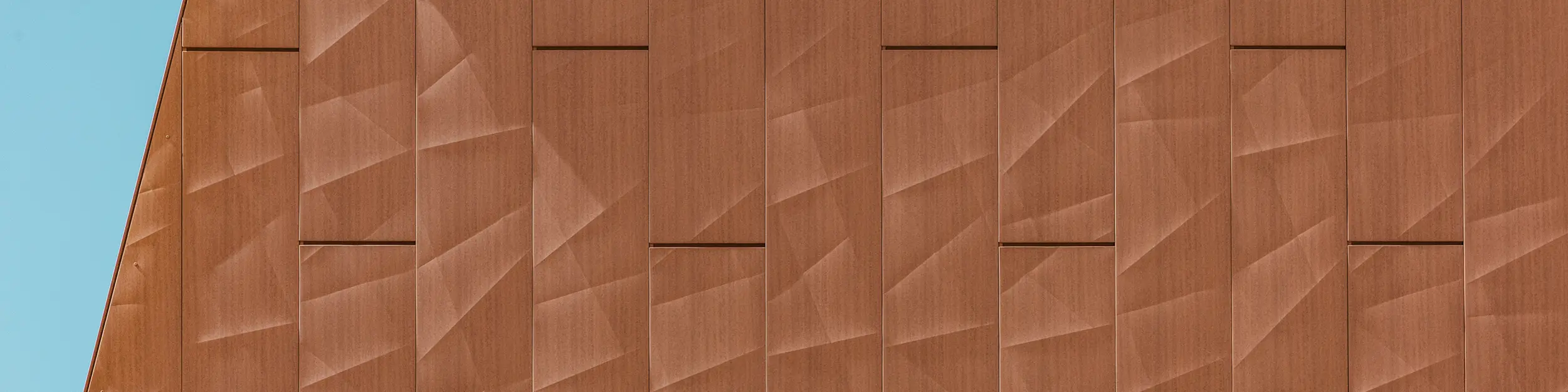 Detailný záber hliníkovej fasády PREFA Siding.X na čerpacej stanici pitnej vody WML v Lottume s precízne ohýbanými panelmi v teplom vzhľade hrdze, ktoré pútajú pozornosť. Geometricky štruktúrovaný povrch vytvára fascinujúcu hru svetla, tieňa a jemných línií, ktorá sa v závislosti od uhla pohľadu živým spôsobom mení. Vysoko kvalitné hliníkové panely PREFA presviedčajú nízkou hmotnosťou, vysokou odolnosťou voči poveternostným vplyvom, stálosťou farieb a úplnou recyklovateľnosťou – pre dlhotrvajúce, udržateľné a nenáročné na údržbu fasádne riešenie.